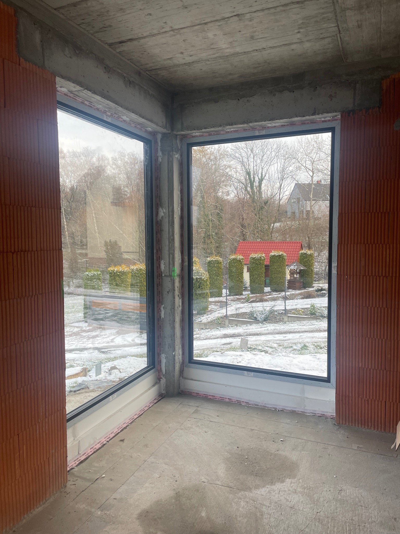 Surowy stan budynku z dużymi oknami z czarnymi ramami, widok na zaśnieżony ogród z czerwoną altaną.