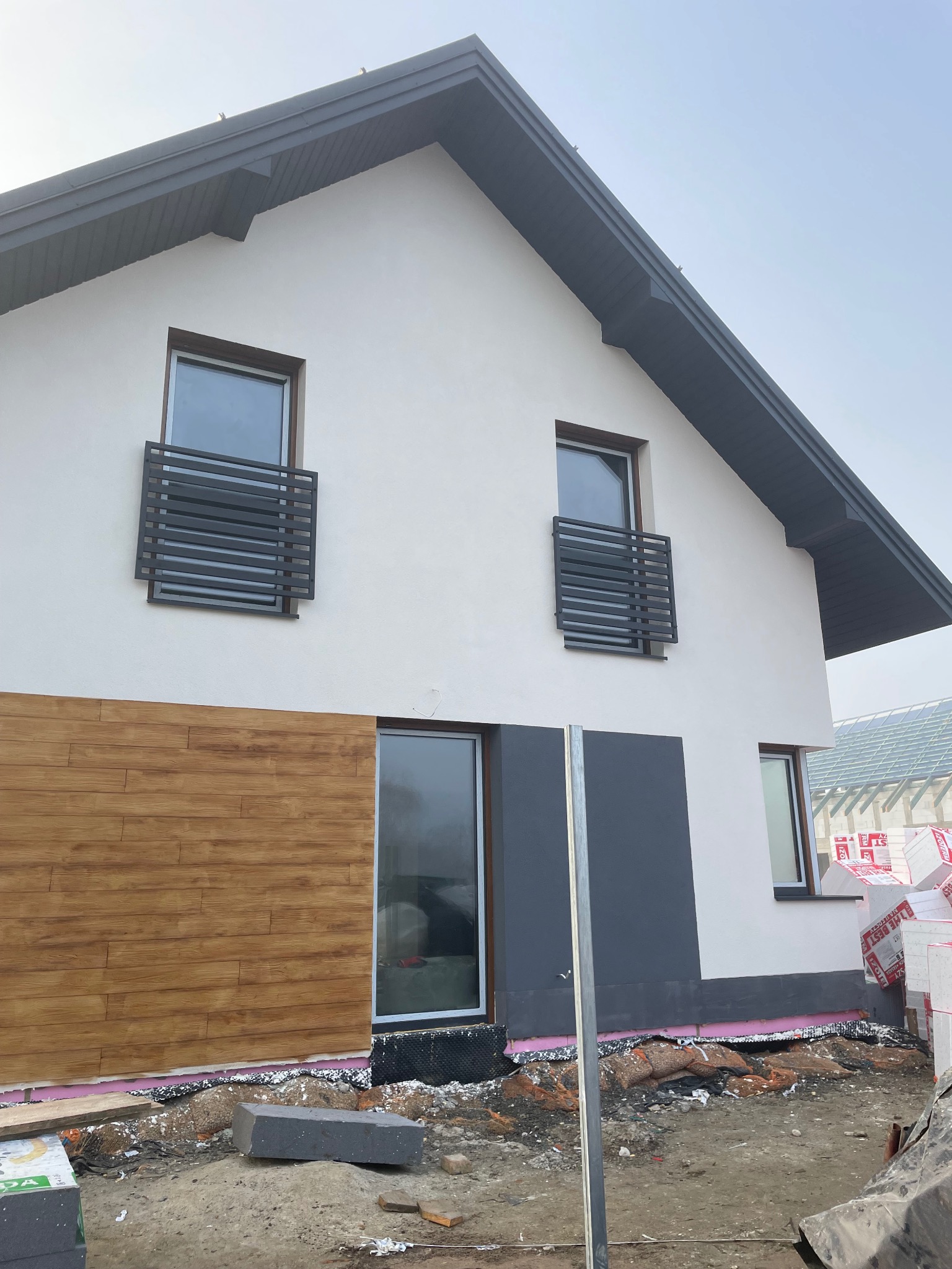 Nowoczesny dom jednorodzinny w trakcie wykończenia elewacji, widoczne okna z ozdobnymi kratkami balkonowymi, fragment drewnianej okładziny elewacyjnej oraz materiały izolacyjne przy fundamencie.