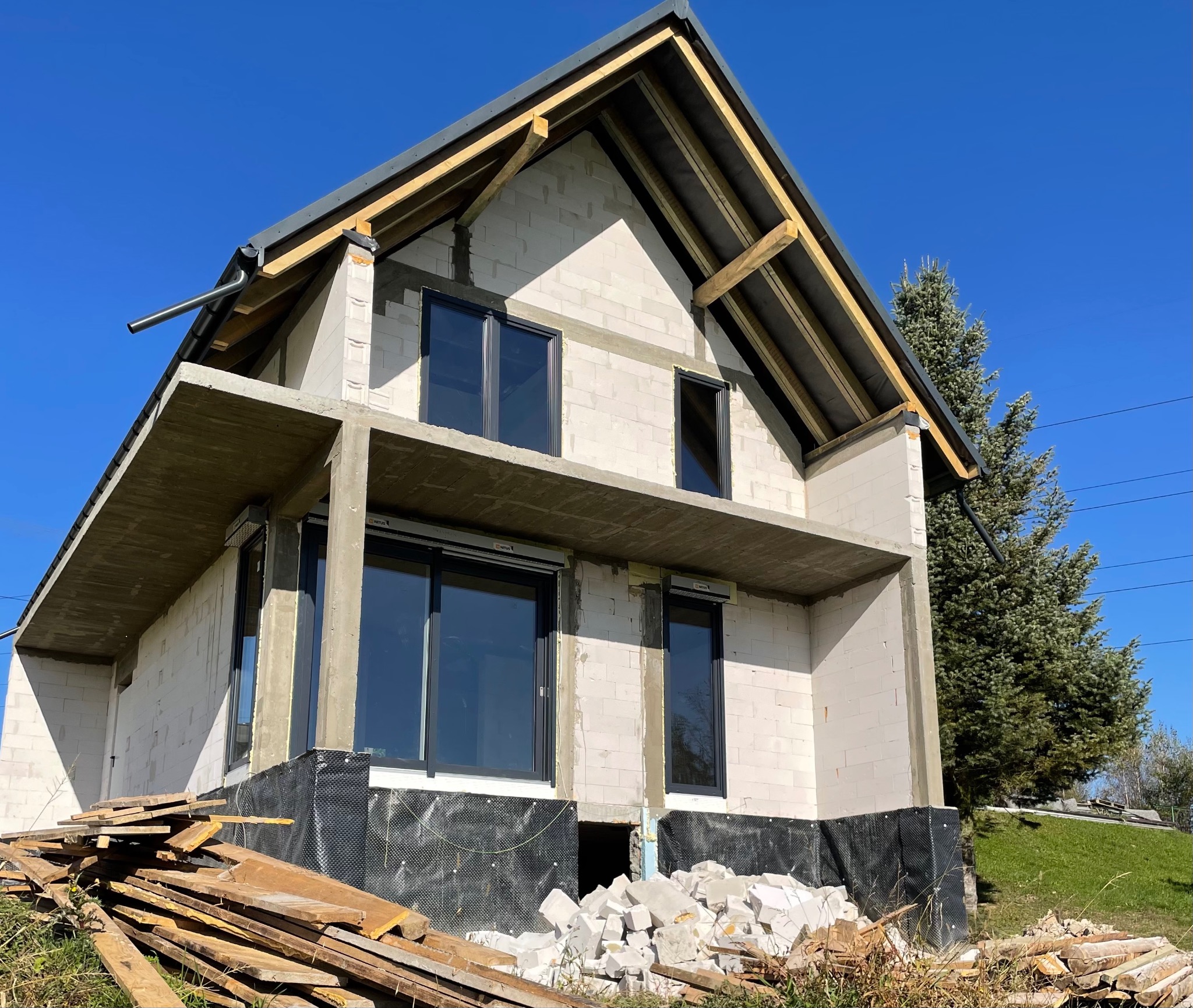 Surowy stan budynku w trakcie budowy, widoczne czarne okna, białe ściany z betonu komórkowego, drewniana konstrukcja dachu, składowane deski i gruz na zewnątrz, błękitne niebo.