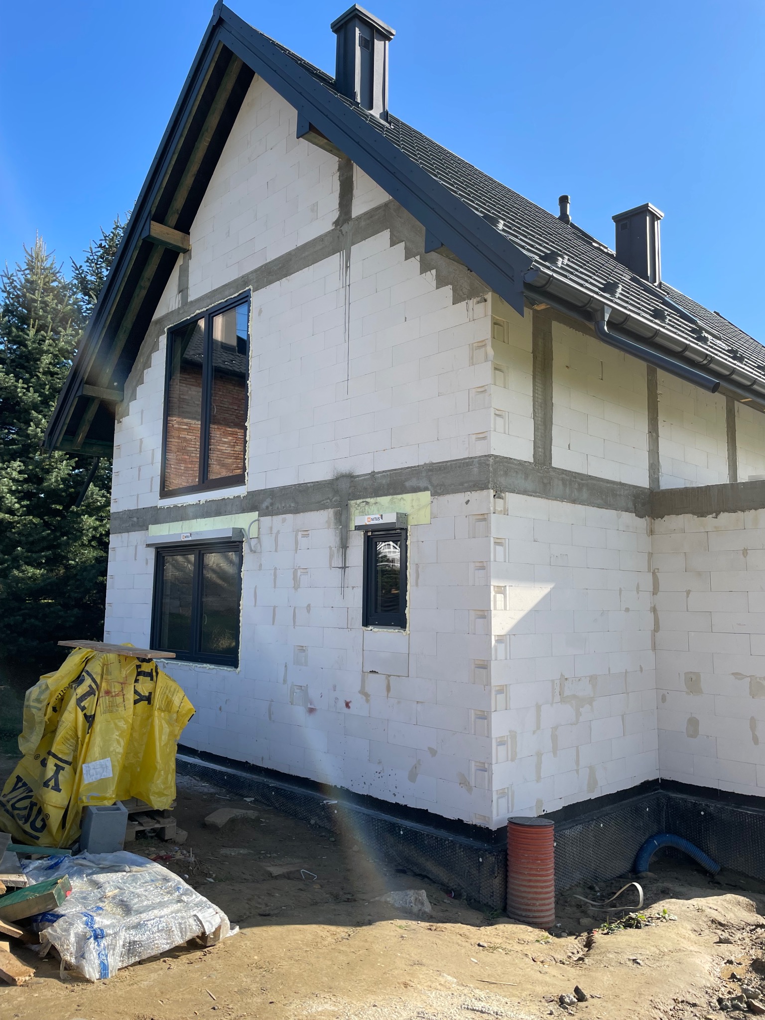 Budowa domu jednorodzinnego w trakcie: wstawione okna z czarnymi ramami, ściany z białych bloczków, czarny dach z kominami, widoczne materiały budowlane i narzędzia.