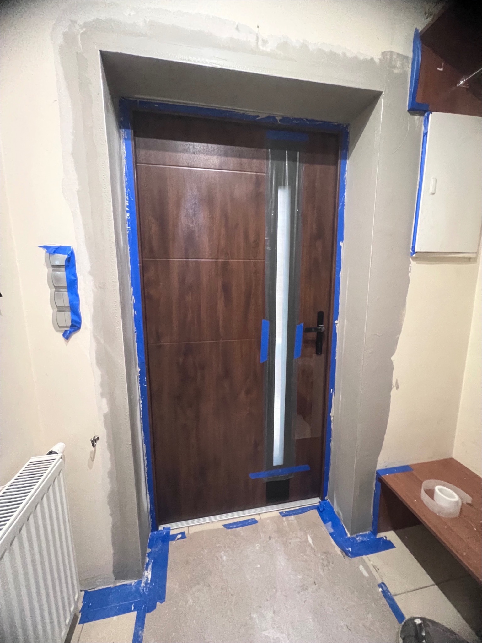 Montaż drzwi wewnętrznych z szybą, zabezpieczonych niebieską taśmą malarską, widoczne ślady po szpachlowaniu ościeżnicy i ściany, grzejnik oraz fragment podłogi zabezpieczone przed zabrudzeniem.