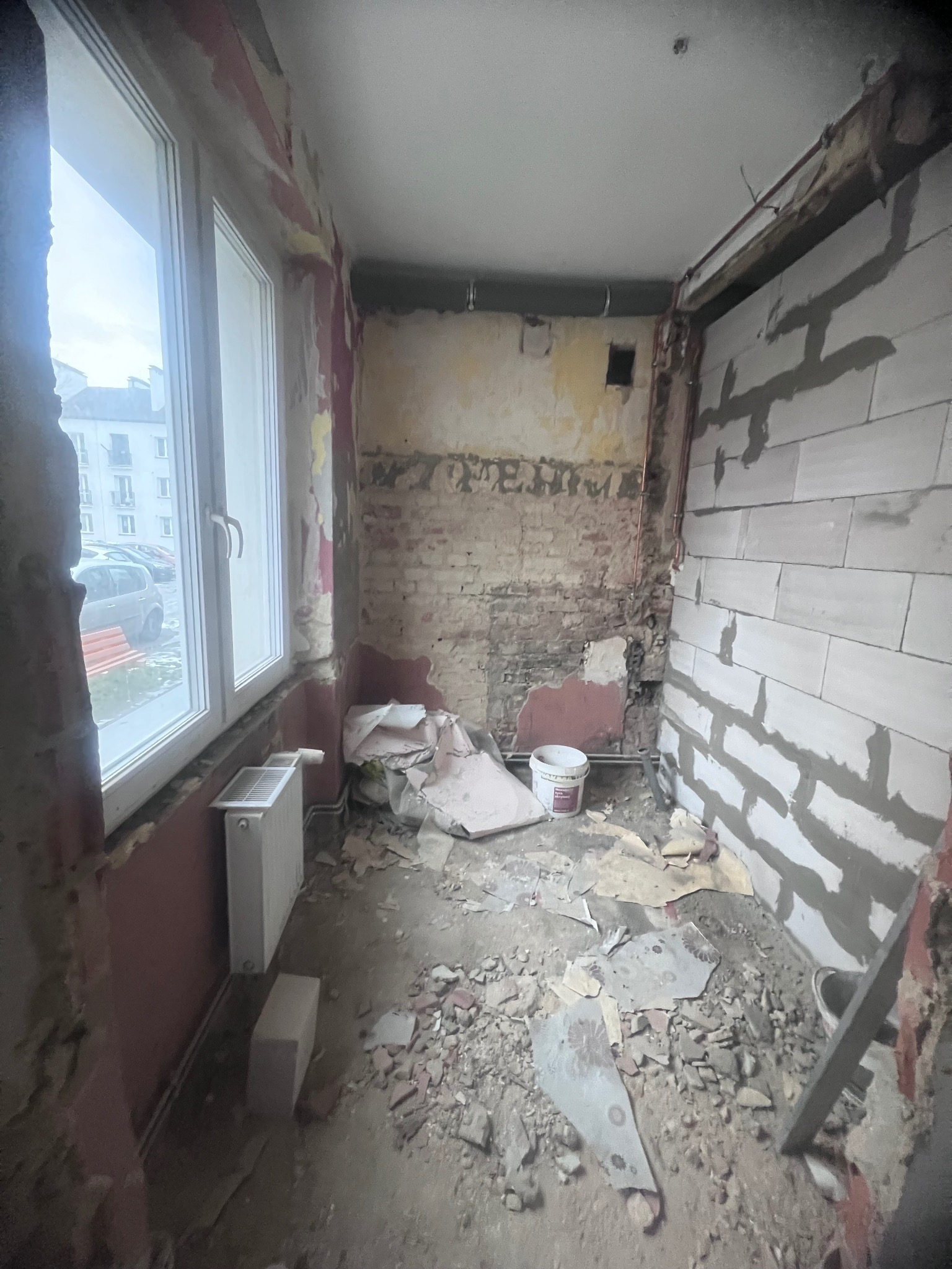 Wnętrze balkonu w trakcie prac remontowych: widoczne fragmenty starej cegły, ślady po zerwanych tapetach, nowe ściany z betonu komórkowego, gruz i resztki materiałów na podłodze, grzejnik pod oknem.
