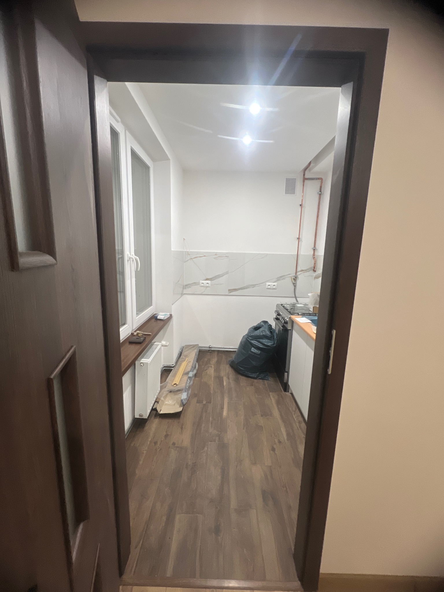 Widok z korytarza na wyremontowaną kuchnię: panele podłogowe, białe ściany z marmurkowym wzorem, okno z brązowym parapetem, grzejnik i czarny worek na śmieci.