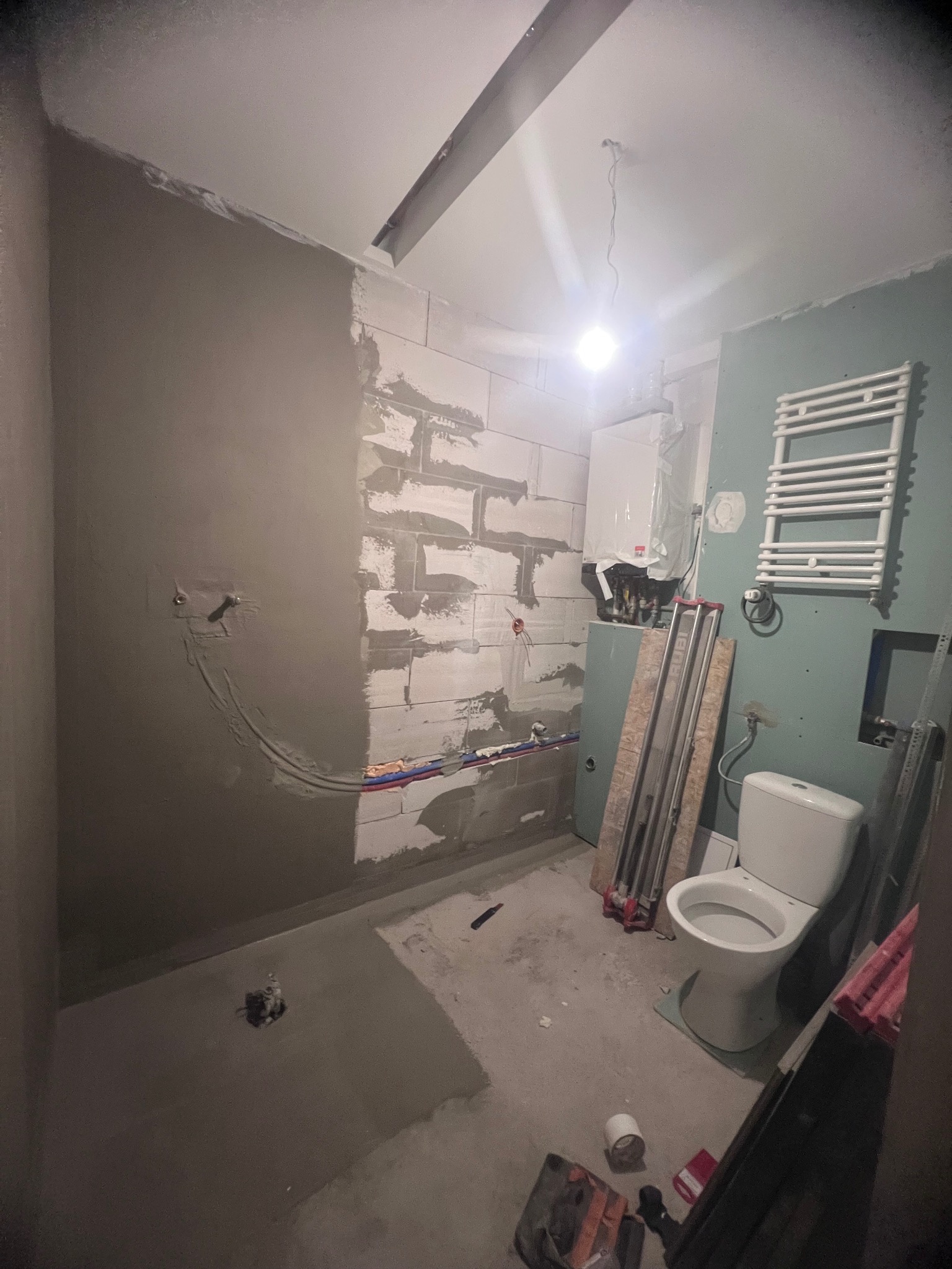 Łazienka w trakcie remontu: ściany częściowo otynkowane, widoczne przewody, grzejnik drabinkowy, biała toaleta, narzędzia i materiały budowlane na podłodze, surowy stan wykończenia.
