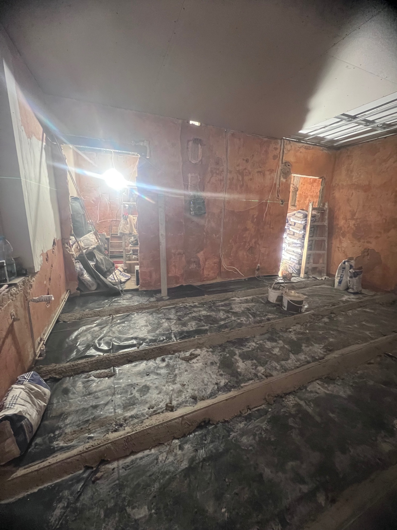 Pokój w trakcie remontu z widocznymi elementami przygotowywanymi pod wylewkę, worki z zaprawą i drabina oparta o ścianę w tle.