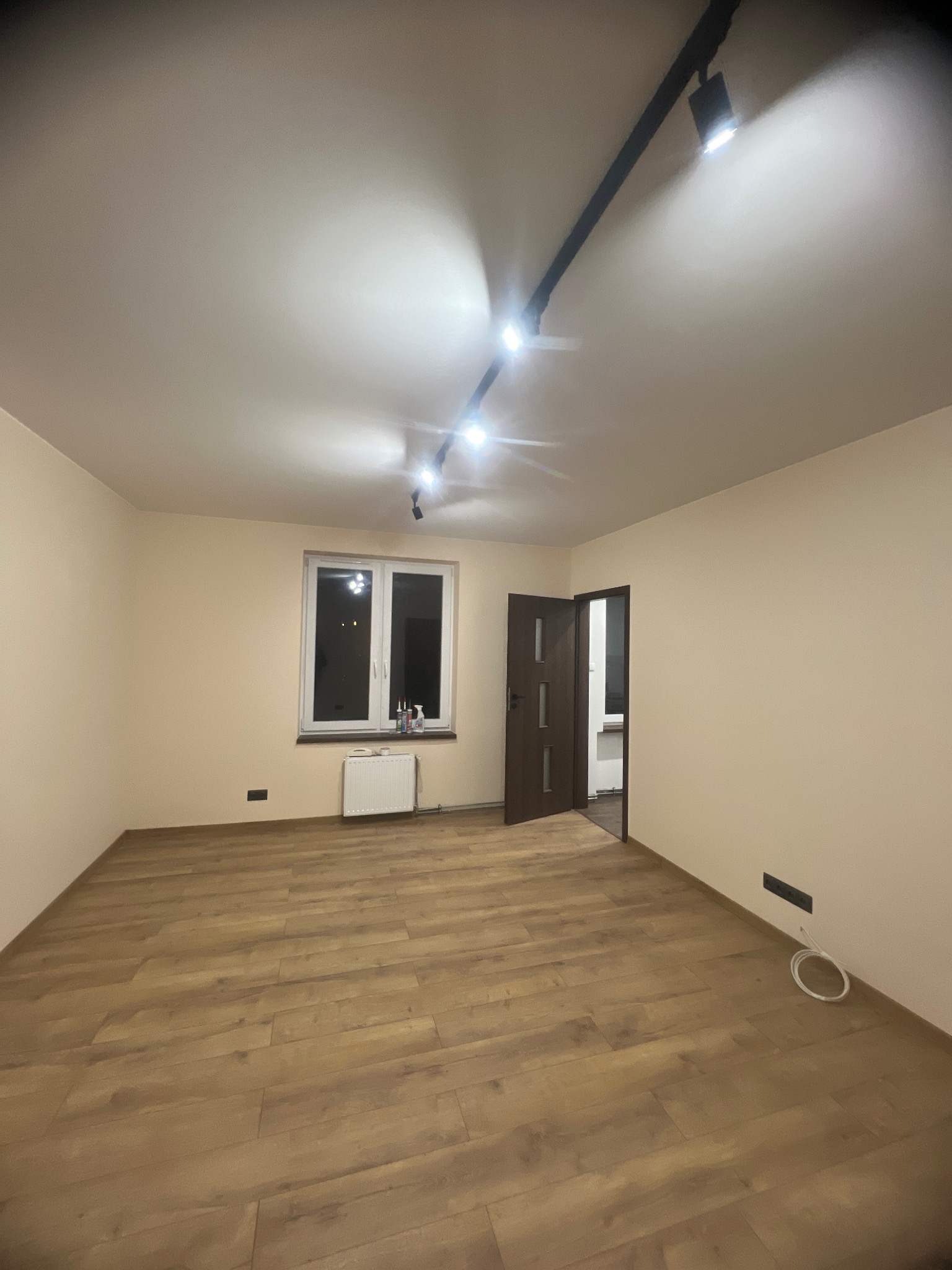 Puste, jasne pomieszczenie po remoncie z panelami podłogowymi, świeżo pomalowanymi ścianami, otwartymi drzwiami i nowoczesnym oświetleniem szynowym na suficie.