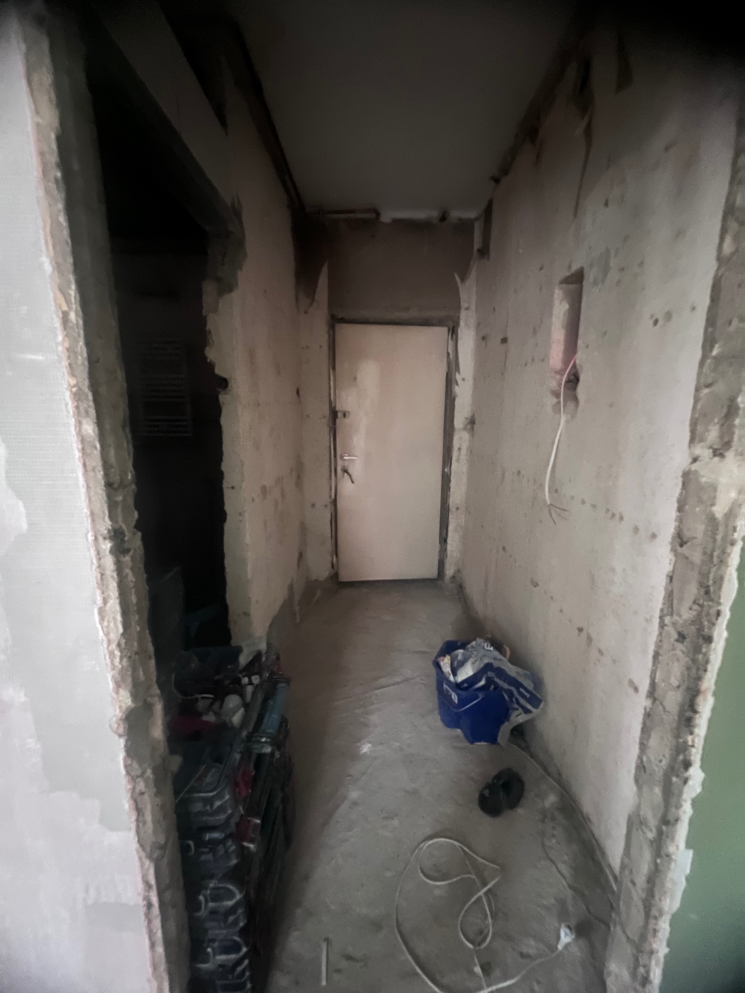 Korytarz w trakcie remontu: surowe ściany z odsłoniętym betonem i resztkami tynku, widoczne przewody elektryczne, worek z gruzem i narzędzia w przejściu do innego pomieszczenia.