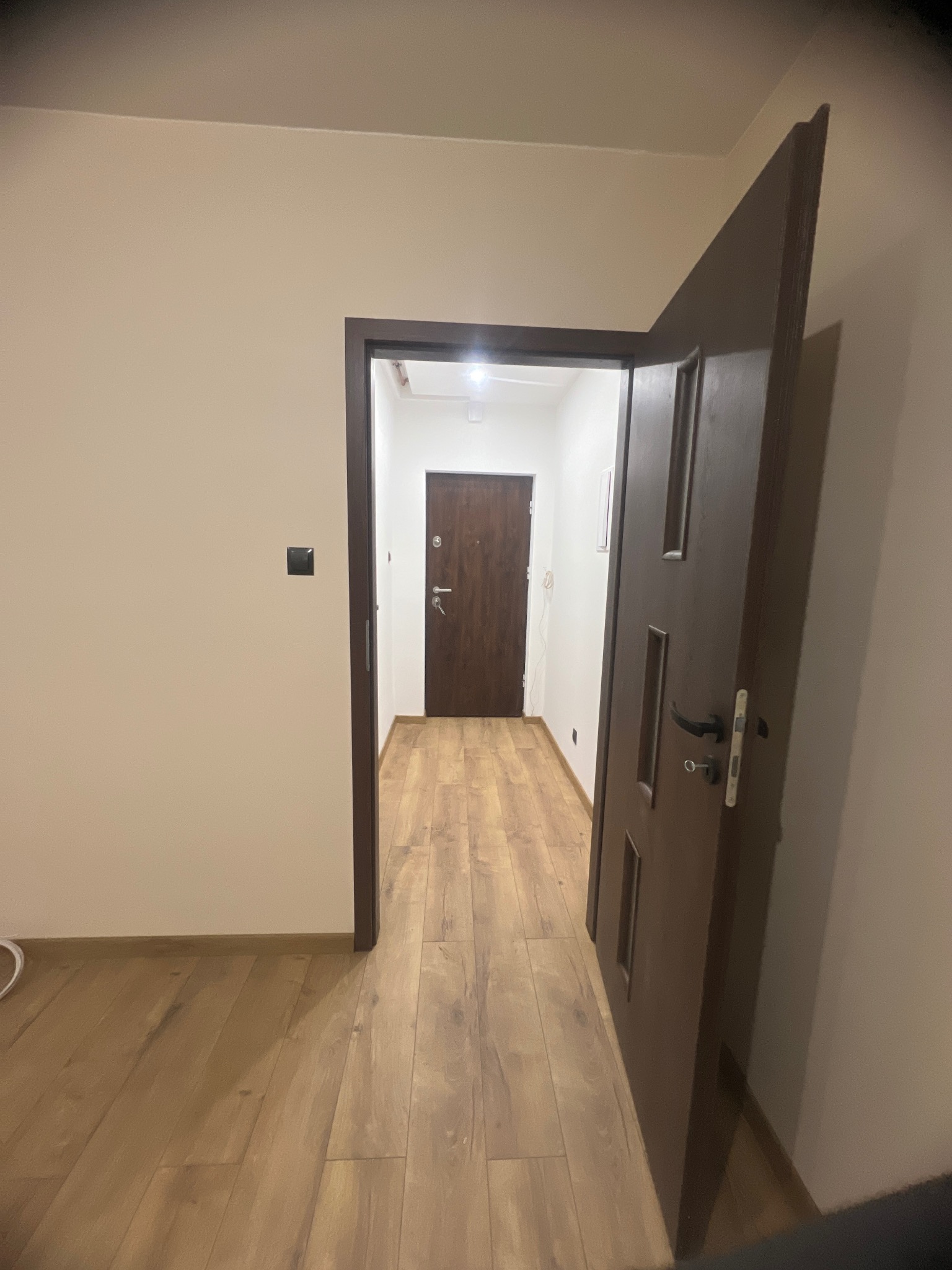 Otwarta, brązowa, wewnętrzna ościeżnica drzwiowa z widocznym kluczem w zamku, prowadząca do korytarza z panelami podłogowymi i brązowymi drzwiami wejściowymi.