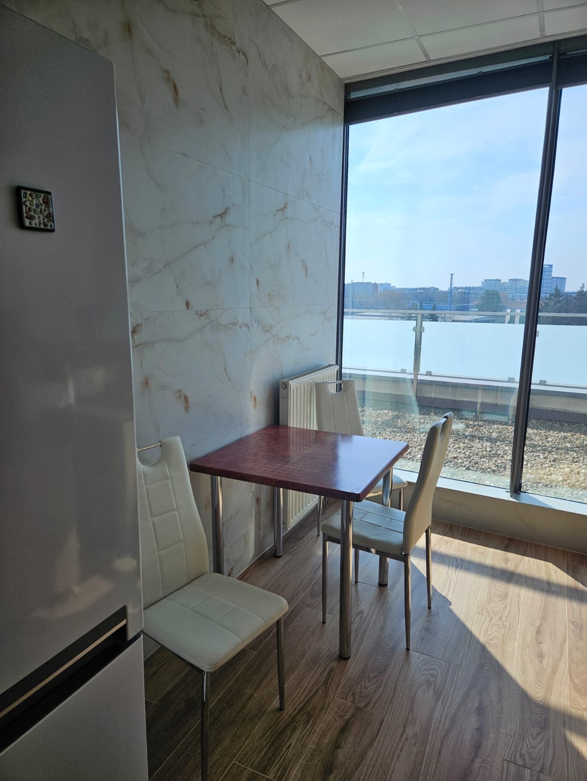 Jasne wnętrze z marmurową ścianą, widokiem na miasto przez duże okno, stół z czerwonym blatem i krzesłami na metalowych nogach, drewnopodobna podłoga, fragment lodówki po lewej.