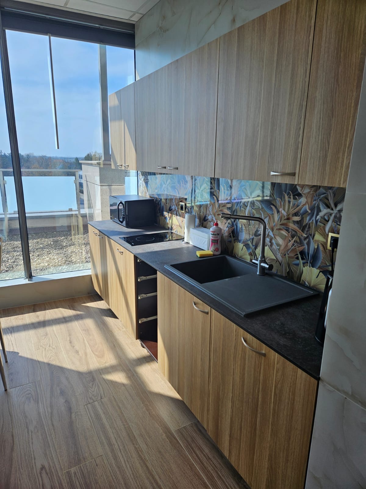 Nowoczesna kuchnia w biurze z drewnianymi szafkami, czarnym blatem i płytkami z motywem liści, widok z okna na panoramę miasta.