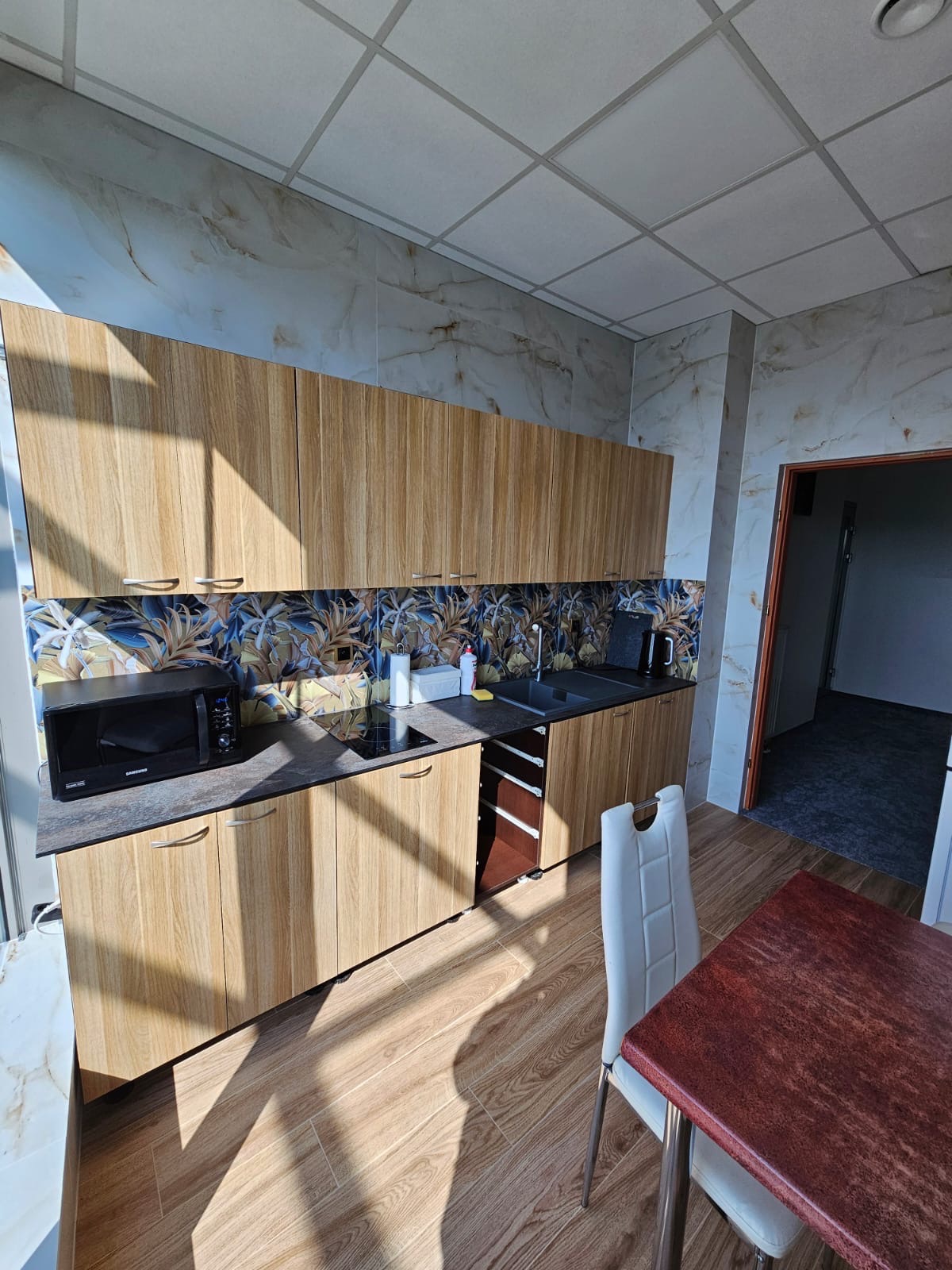 Jasna kuchnia z drewnianymi szafkami, wzorzystym backsplashiem i marmurowymi płytkami ściennymi, oświetlona naturalnym światłem wpadającym przez okno.