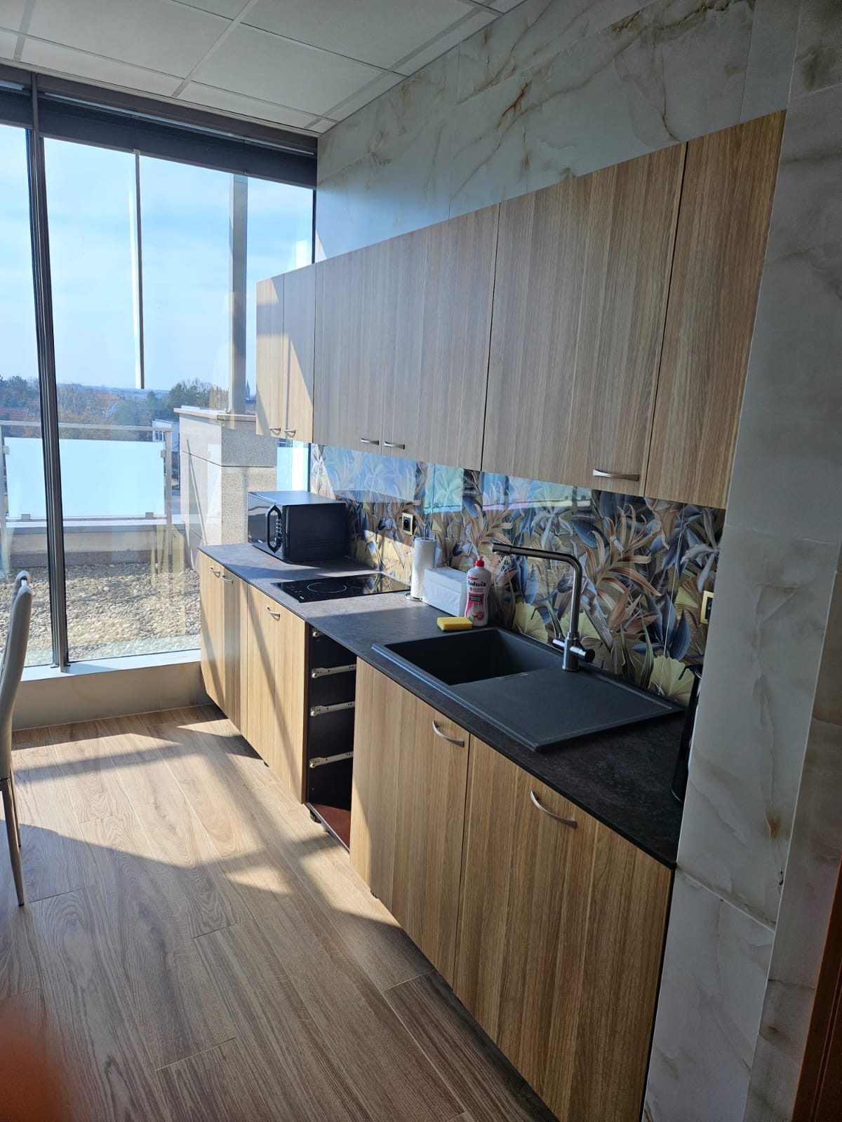 Jasna kuchnia z drewnianymi szafkami i blatem w kolorze grafitu, z widokiem na miasto przez panoramiczne okno, z wzorzystą ścianą nad zlewem.