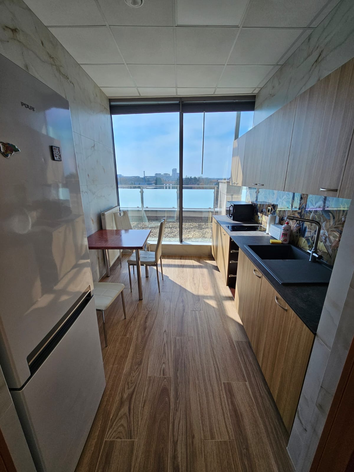 Wnętrze kuchni z drewnianymi panelami podłogowymi i zabudową kuchenną, widok na miasto z balkonu przez duże okno, marmurowe wykończenie ścian.