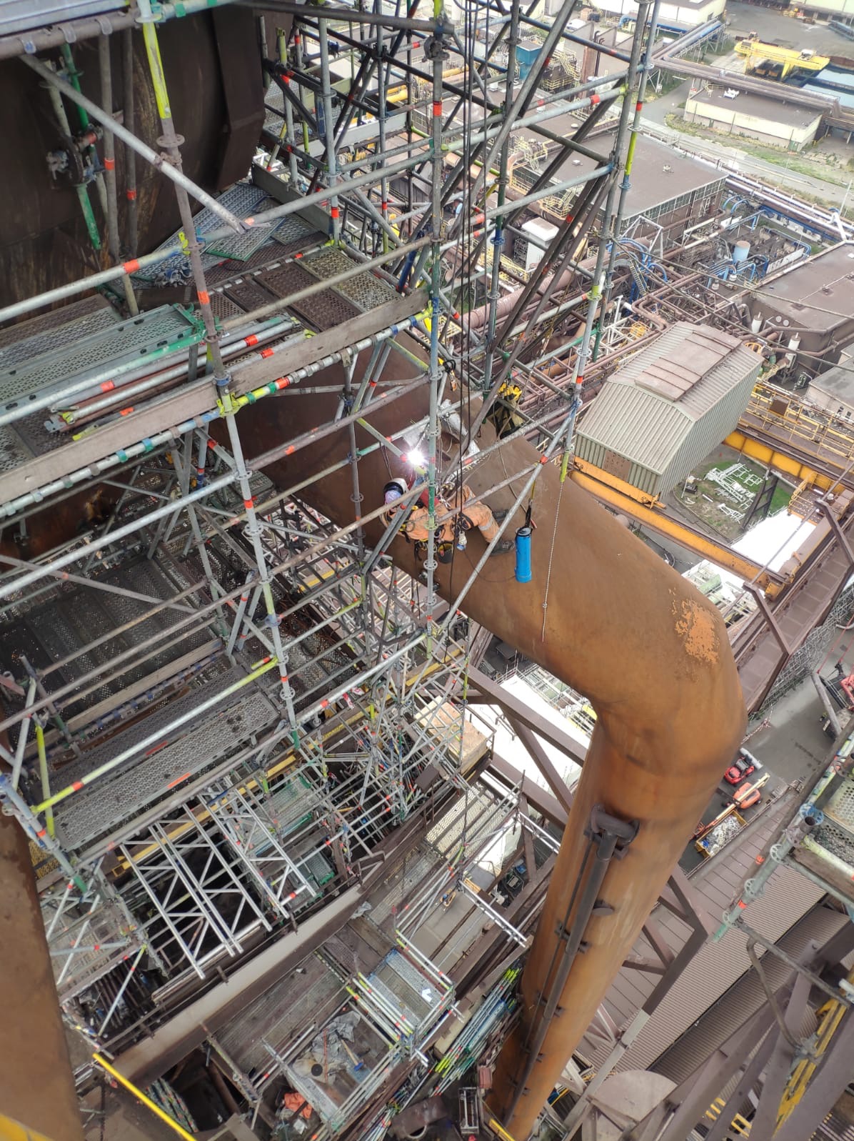 Spawacz w pomarańczowym kombinezonie i kasku, pracujący na wysokościach na rusztowaniu, spawający grubą, rdzawą rurę w hucie. Widoczny błysk spawarki i butla z gazem obok spawacza.