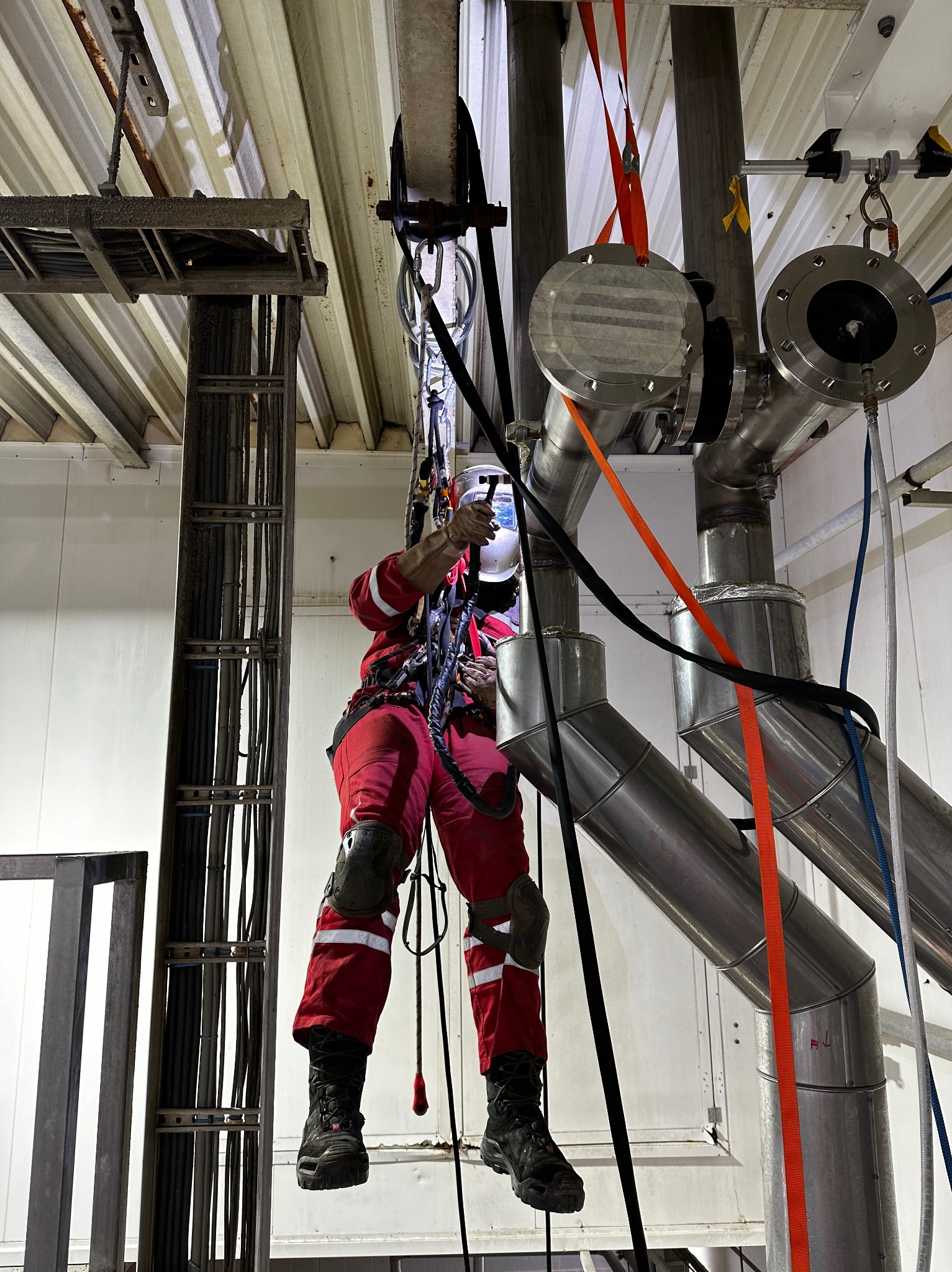 Spawacz w czerwonym kombinezonie, zabezpieczony linami, wykonuje prace spawalnicze na rurociągu ze stali nierdzewnej w hali produkcyjnej. Widoczne elementy instalacji, kable oraz konstrukcja...