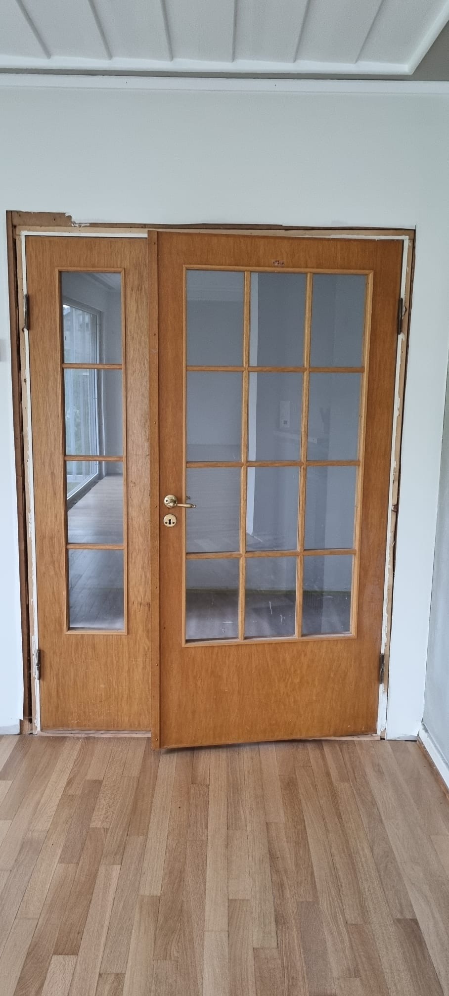 Drewniane, przeszklone drzwi dwuskrzydłowe w trakcie montażu, widoczne niedokończone obramowanie i szczeliny przy ścianie.