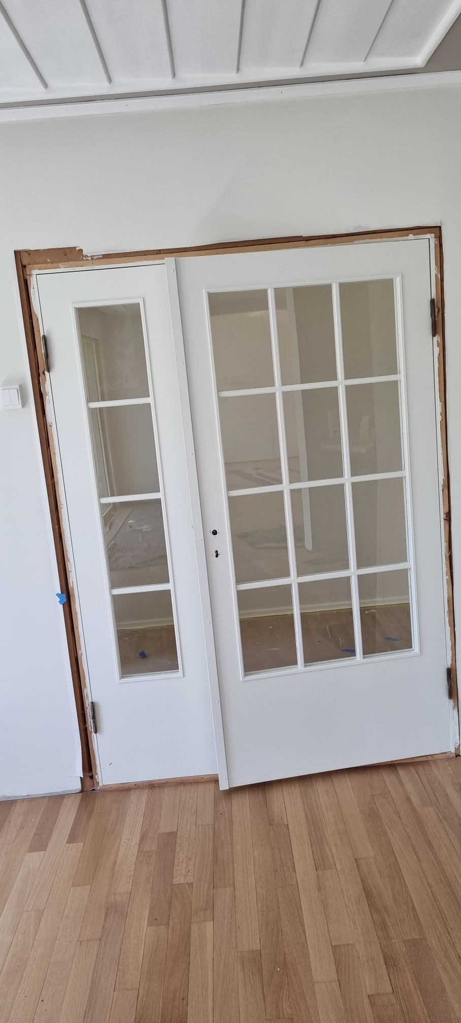 Montaż dwuskrzydłowych, białych drzwi z szybami w nowym otworze, widoczne surowe drewno ościeżnicy i ściana po obróbce, zabezpieczona podłoga.