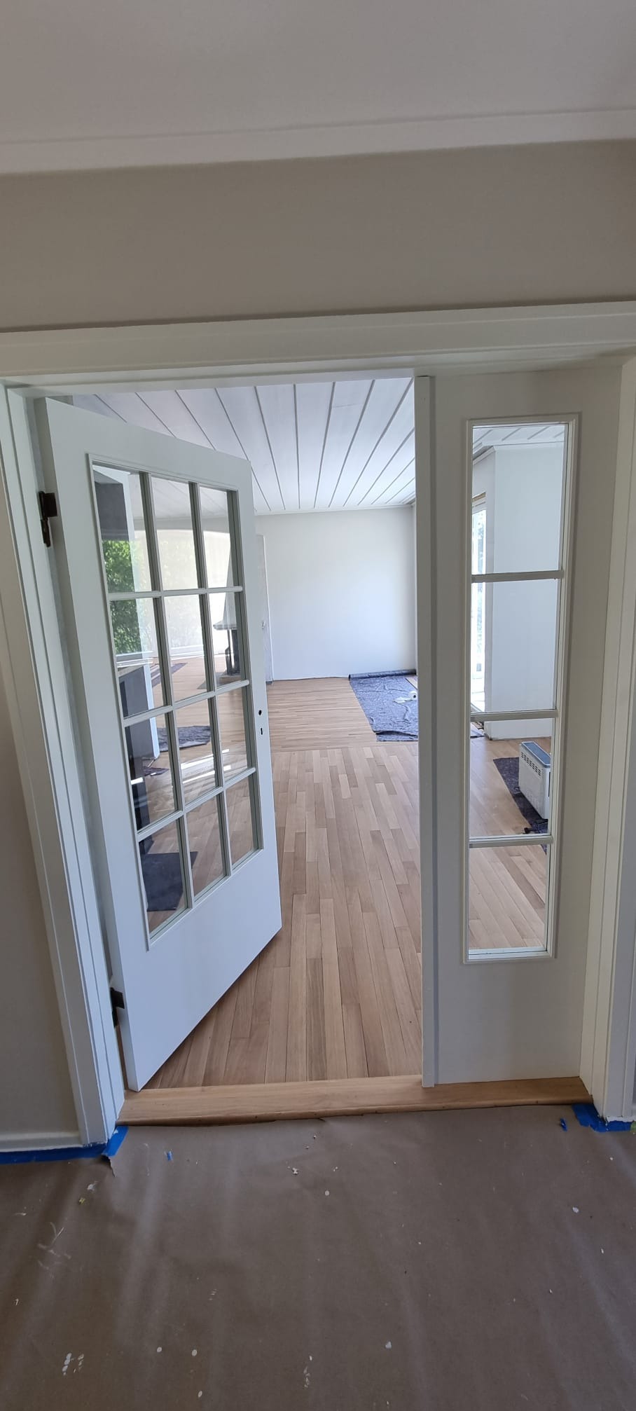 Widok na odnowione wnętrze z parkietem i pomalowanymi ścianami przez otwarte, białe, przeszklone drzwi. Podłoga zabezpieczona papierem malarskim i taśmą.