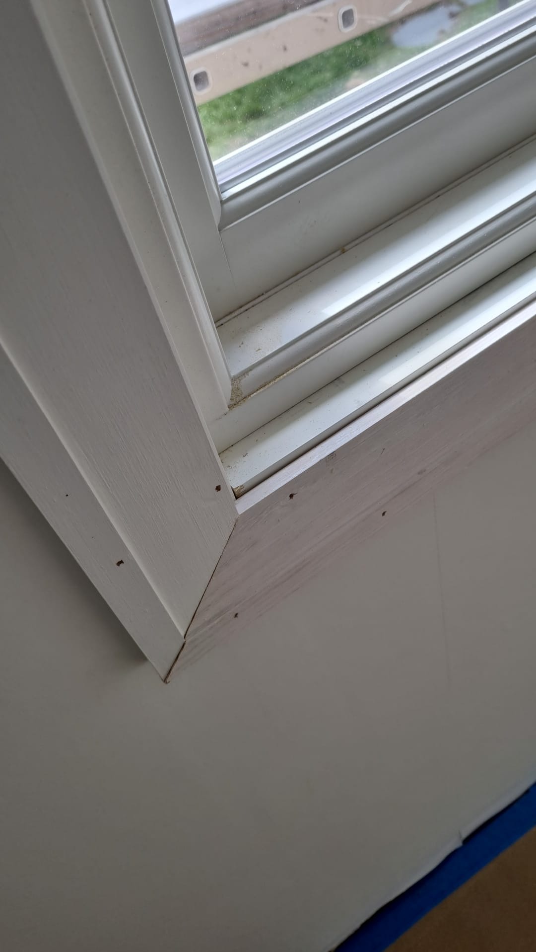 Fragment okna z drewnianą ramą w trakcie malowania na biało, widoczne ślady po gwoździach i niedomalowane fragmenty, zabezpieczona taśmą malarską ściana.