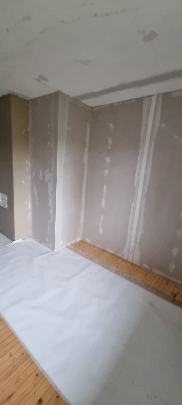 Pokój w trakcie prac, ściany pokryte płytami kartonowo-gipsowymi, widoczne łączenia płyt, podłoga zabezpieczona folią malarską, listwa przypodłogowa częściowo zdjęta.
