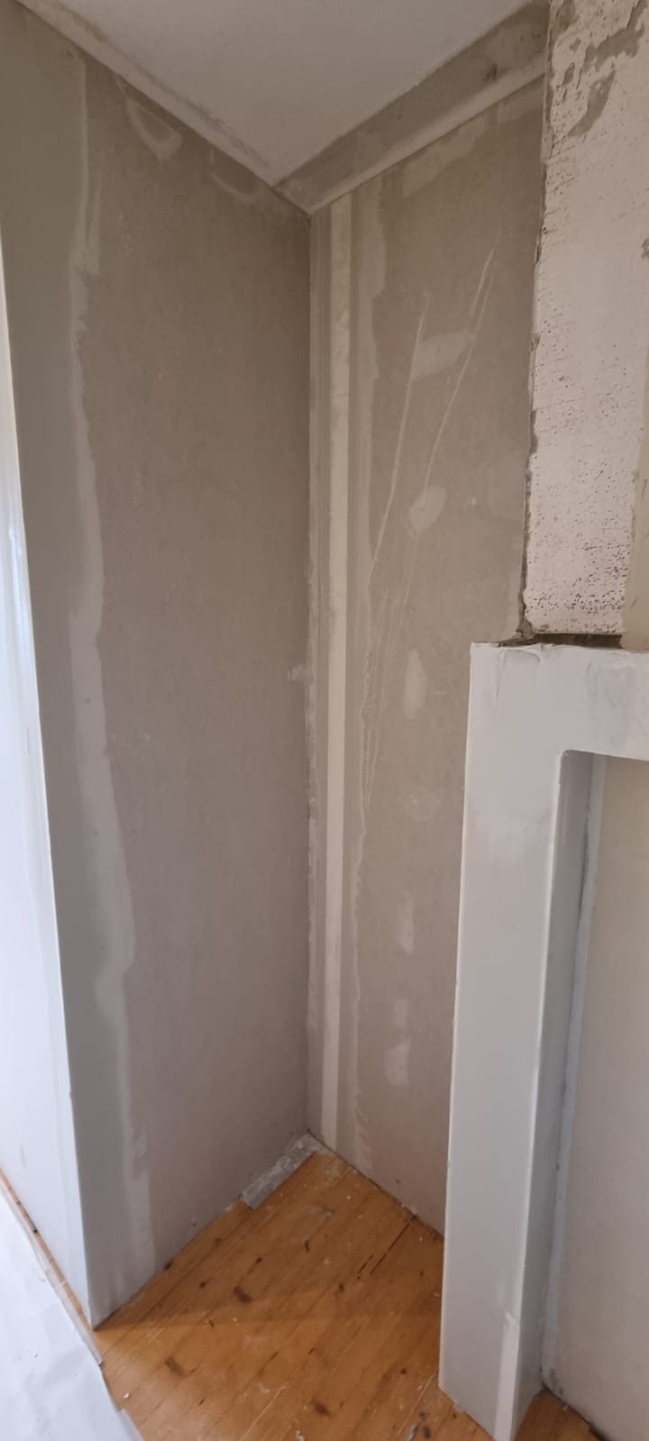 Ściana w rogu pomieszczenia po szpachlowaniu, widoczne nierówności i ślady po wałku, przygotowana do malowania, z fragmentem drewnianej podłogi i białym obramowaniem.