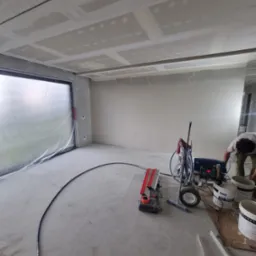 Wnętrze w trakcie prac wykończeniowych: ściany i sufit po nałożeniu gładzi, widoczna maszyna natryskowa do gładzi i zakryte folią okno.