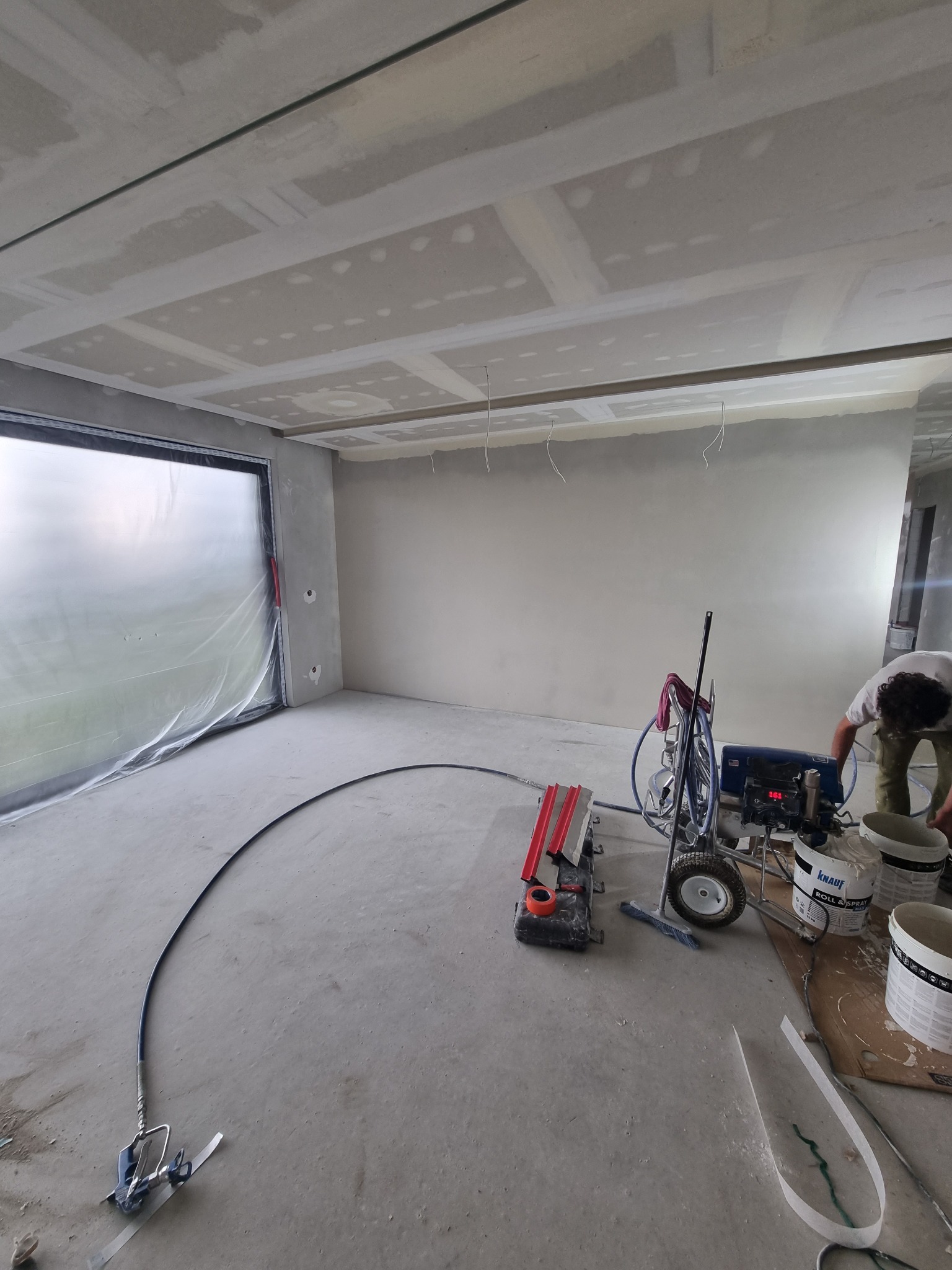 Wnętrze w trakcie prac wykończeniowych: ściany i sufit po nałożeniu gładzi, widoczna maszyna natryskowa do gładzi i zakryte folią okno.