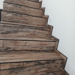 S-build - Schody wyłożone płytkami imitującymi drewno z ciemnymi listwami, widok z dołu, ściana pomalowana na biało.