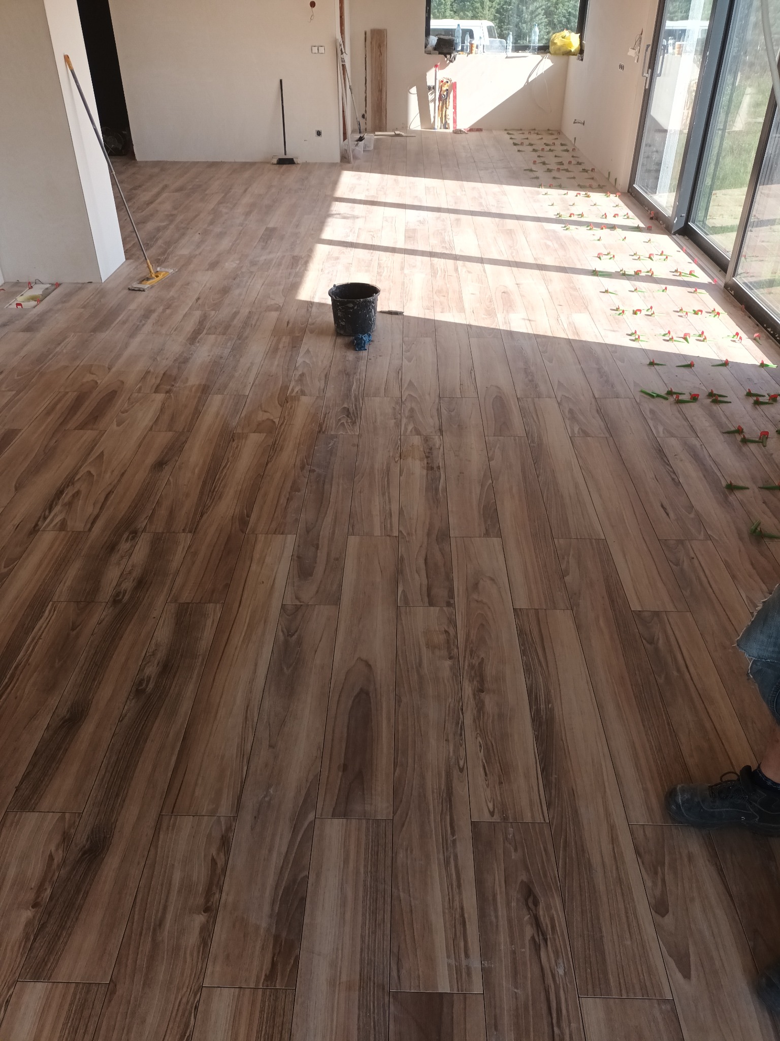 Układanie podłogi imitującej drewno w jasnym pomieszczeniu z dużymi oknami, widoczne kliny dystansowe i wiadro z zaprawą.