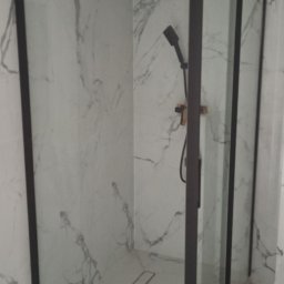 TL TARAS LITSEVYCH - Kabina prysznicowa z czarnymi profilami i marmurowymi płytkami, widoczny odpływ liniowy i słuchawka prysznicowa z miedzianymi elementami, surowa żarówka na kablu.