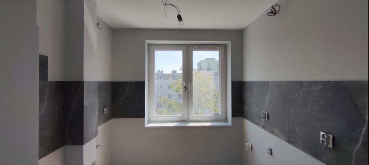 Częściowo wykończona kuchnia z szarymi płytkami imitującymi marmur na ścianach, widoczne gniazdka elektryczne i okno z widokiem na budynek mieszkalny.