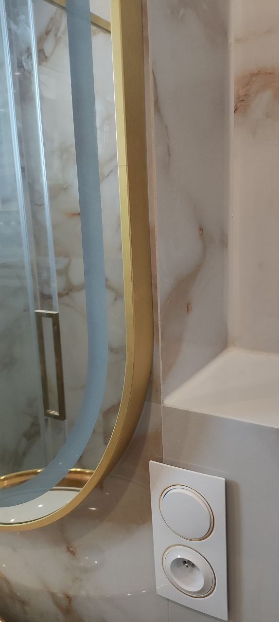 Fragment łazienki z marmurową okładziną ściany, okrągłym lustrem w złotej ramie i białym gniazdkiem elektrycznym z wykończeniem w kolorze złota.