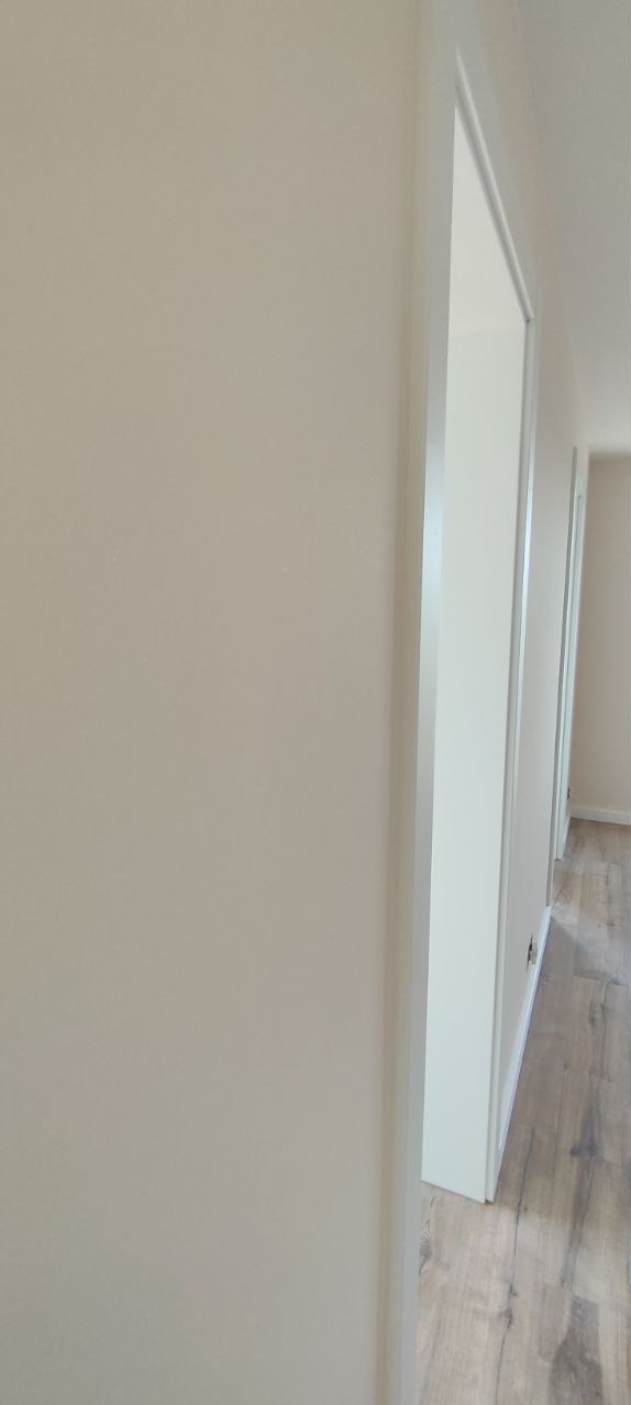 Świeżo pomalowana ściana w odcieniach beżu, widoczna rama białych drzwi i fragment podłogi z paneli w tle