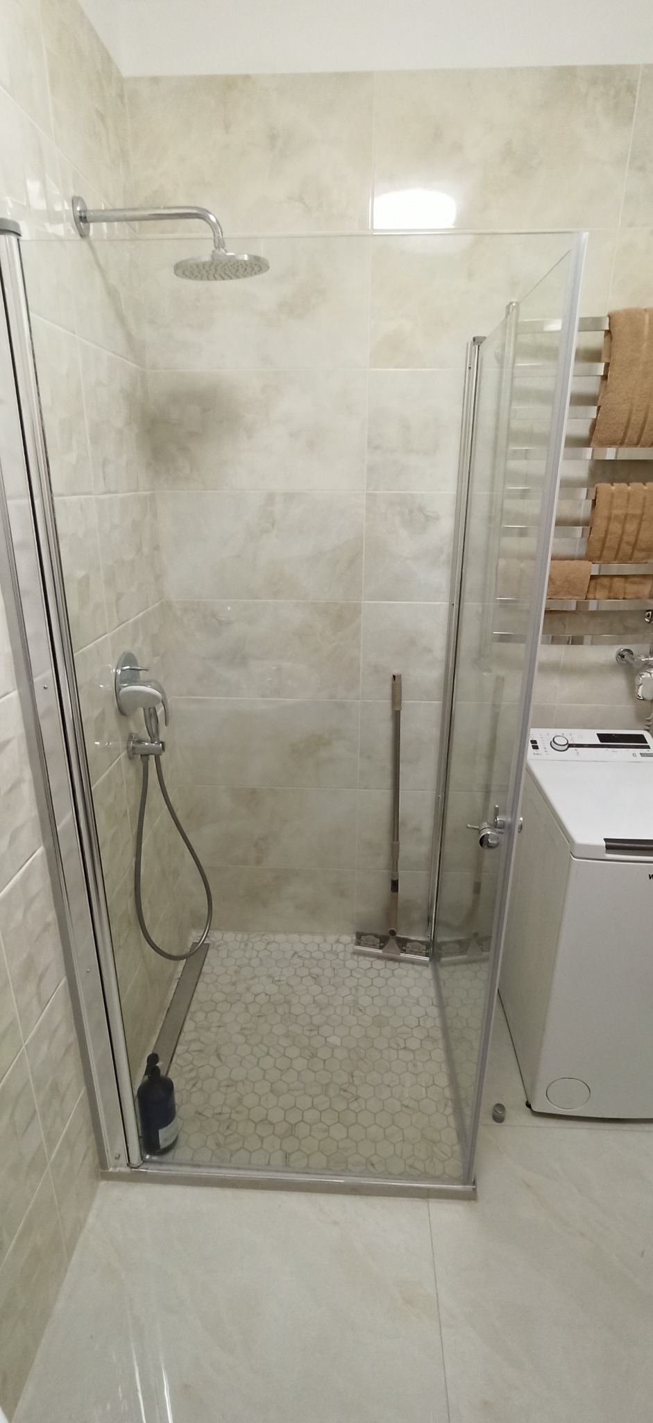 Nowoczesna kabina prysznicowa z deszczownicą, wykończona beżowymi płytkami imitującymi marmur i mozaiką heksagonalną na podłodze, widoczna pralka i grzejnik drabinkowy.