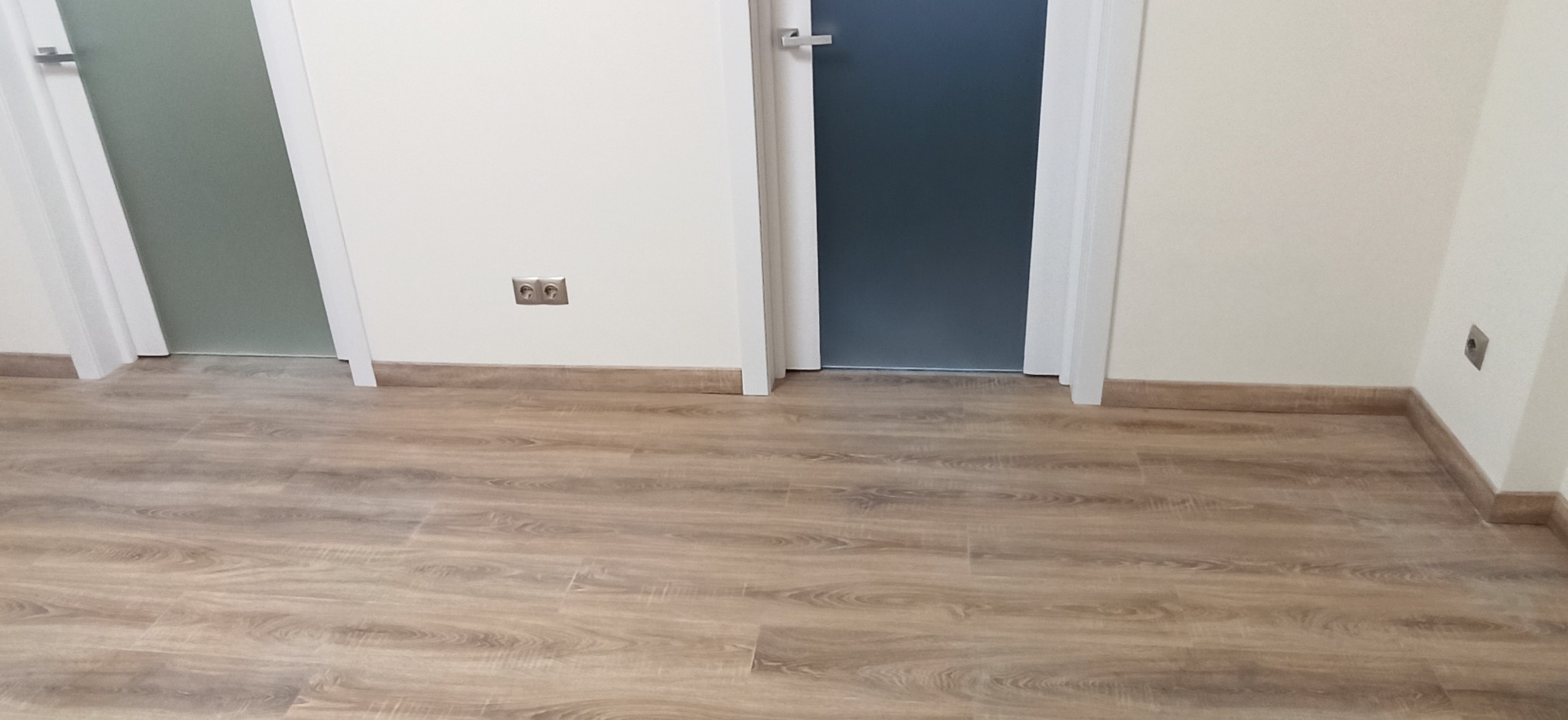 Ułożone panele podłogowe w jasnym odcieniu drewna, widoczne dwie pary drzwi z ościeżnicami, gniazdka elektryczne na ścianie, listwy przypodłogowe w kolorze podłogi.