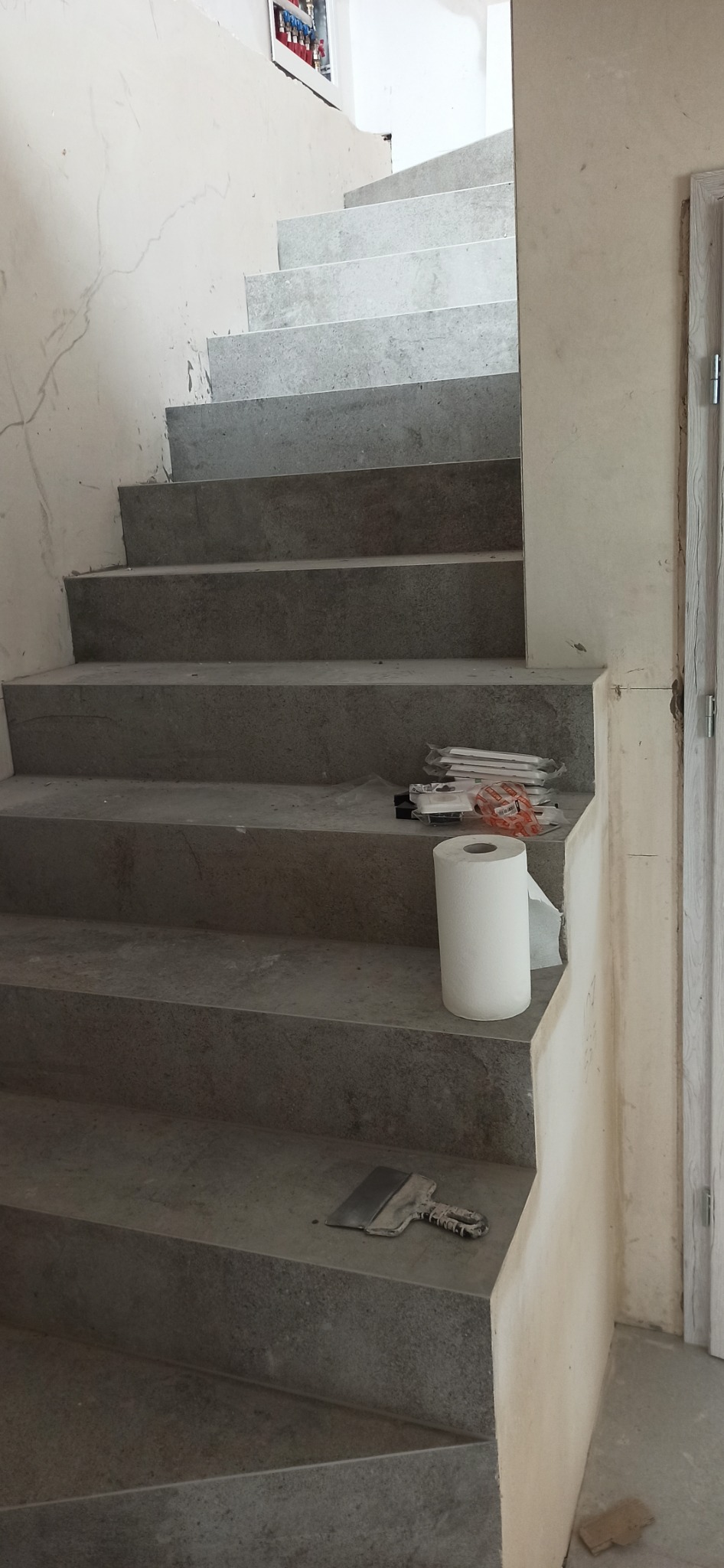 Schody wykończone szarym kamieniem lub płytkami, z widocznymi narzędziami i materiałami budowlanymi, takimi jak szpachelka i rolka papieru, ściany w trakcie prac wykończeniowych.
