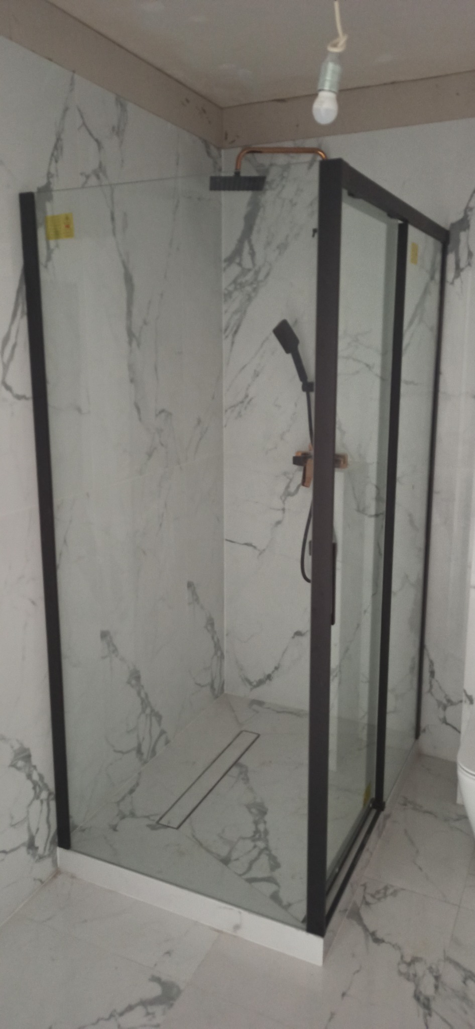Kabina prysznicowa z czarnymi profilami i marmurowymi płytkami, widoczny odpływ liniowy i słuchawka prysznicowa z miedzianymi elementami, surowa żarówka na kablu.