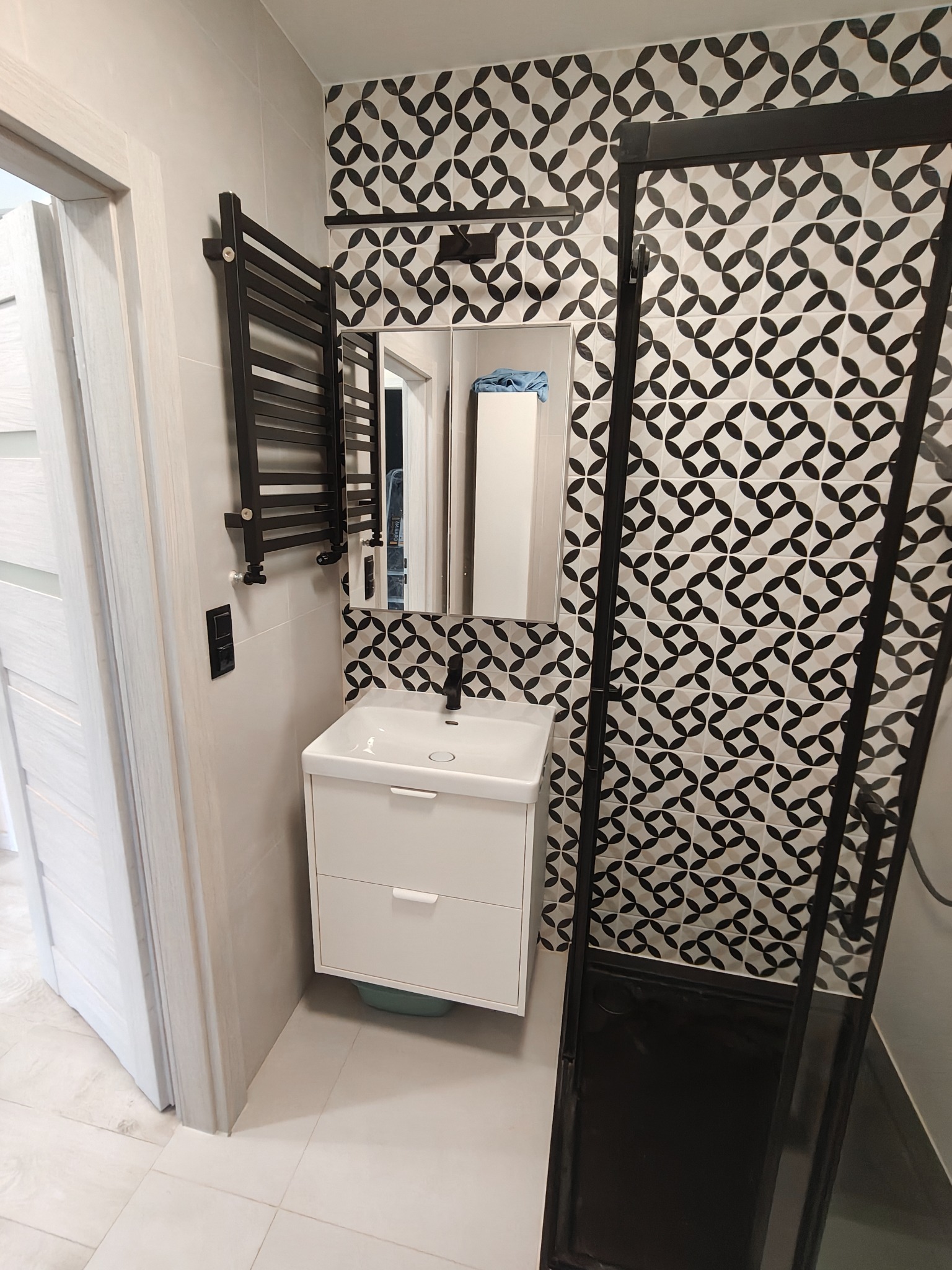 Wykończona łazienka z geometryczną mozaiką na ścianie, białą szafką umywalkową i czarnym grzejnikiem. Kabina prysznicowa z czarną ramą. Ujęcie z perspektywy wejścia.