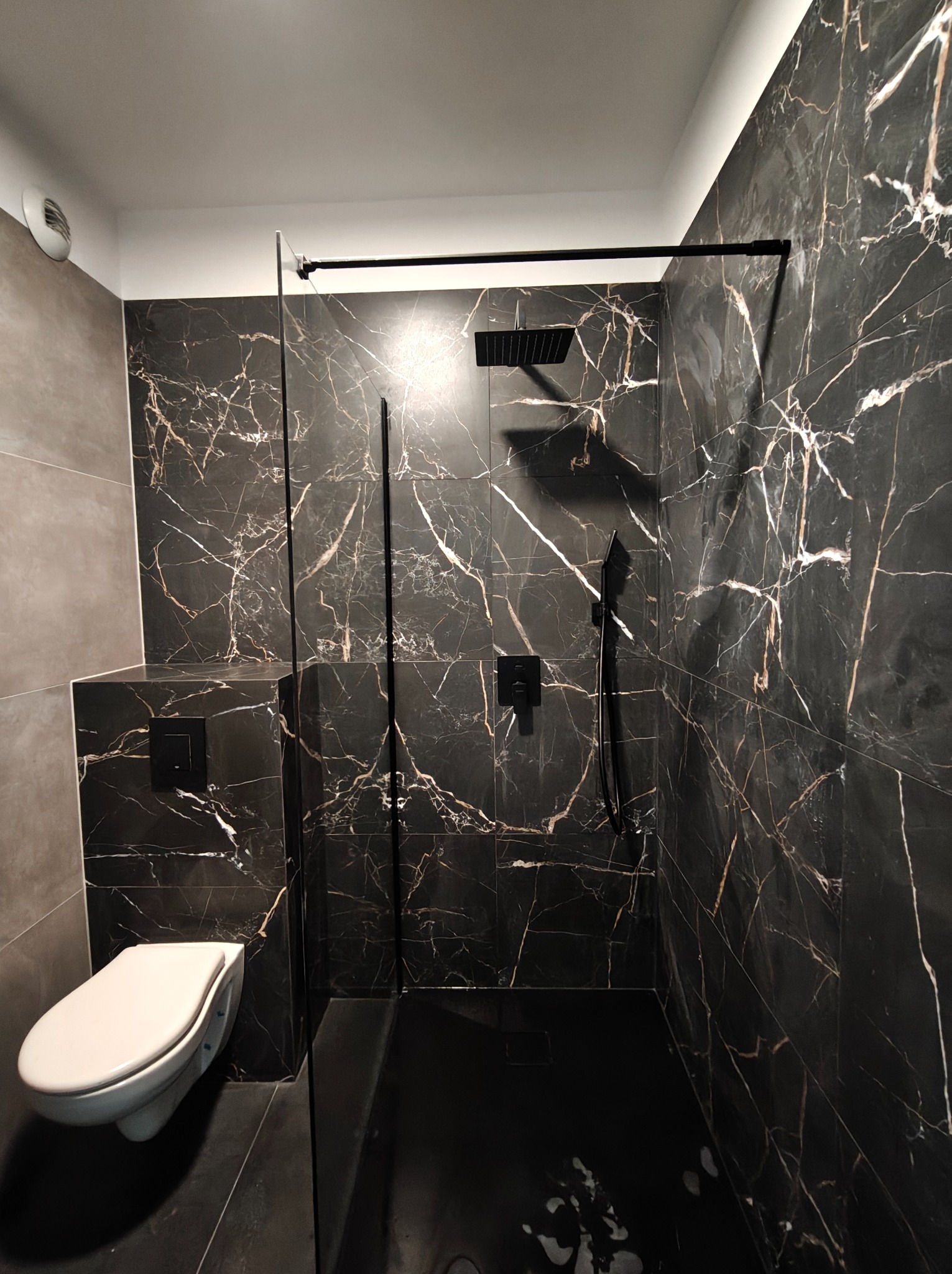 Elegancka łazienka z kabiną typu walk-in, wykończona czarnym marmurem z białymi żyłami i czarną armaturą. Minimalistyczny design z podtynkową spłuczką.