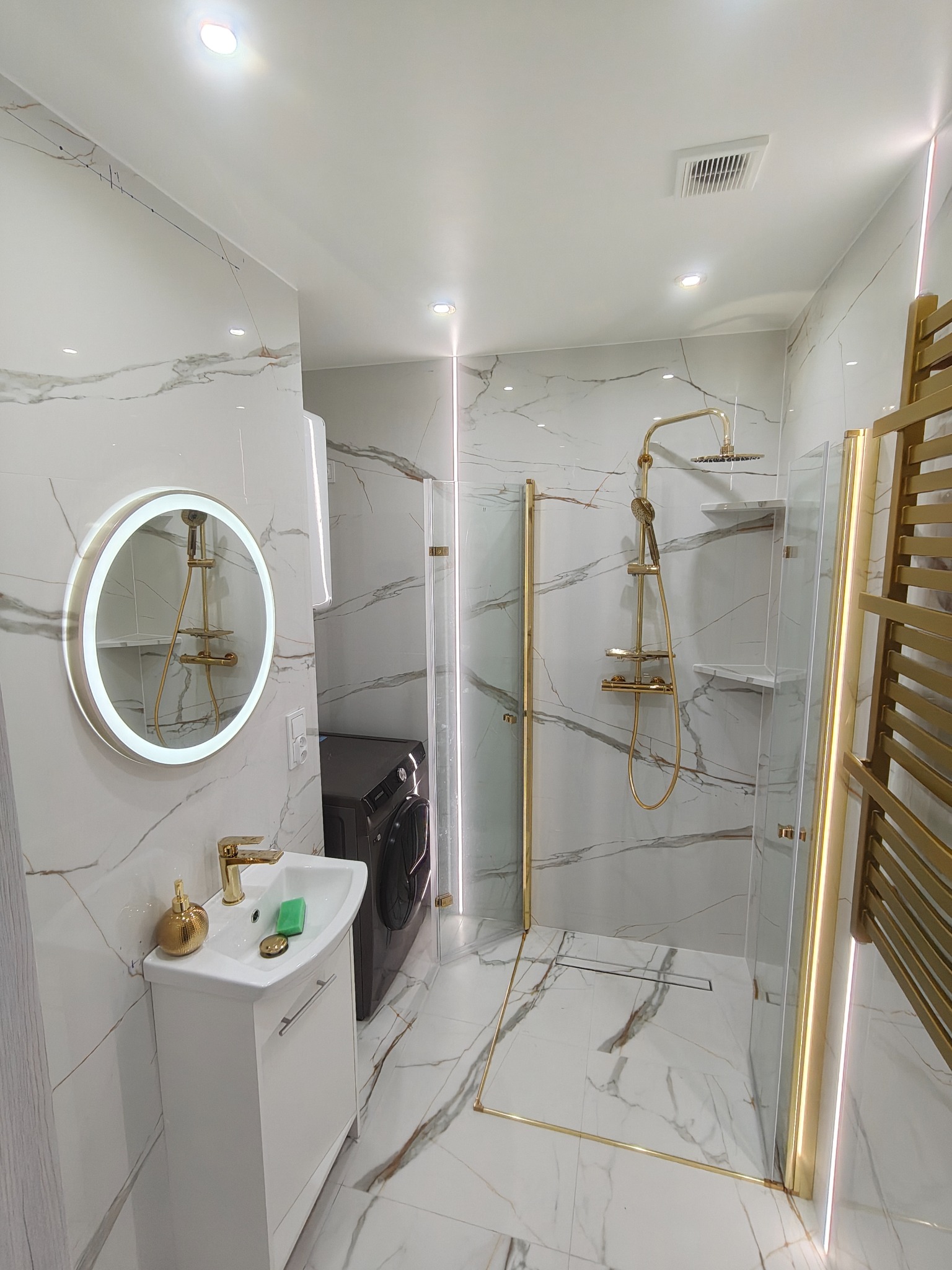 Elegancka łazienka po remoncie: białe marmurowe płytki, złote akcenty armatury, okrągłe lustro z podświetleniem i kabina prysznicowa ze złotymi ramami.