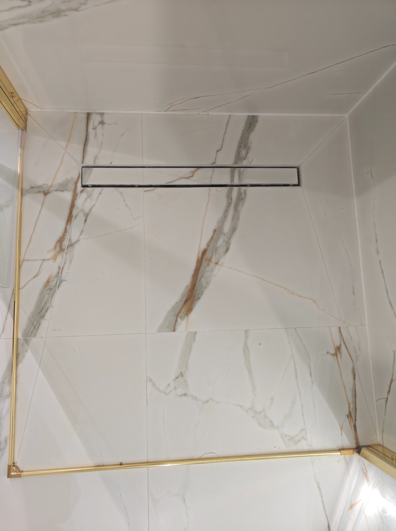 Wykończona kabina prysznicowa z odpływem liniowym i białymi płytkami imitującymi marmur, złote obramowanie kabiny, nowoczesny design, widok z góry.