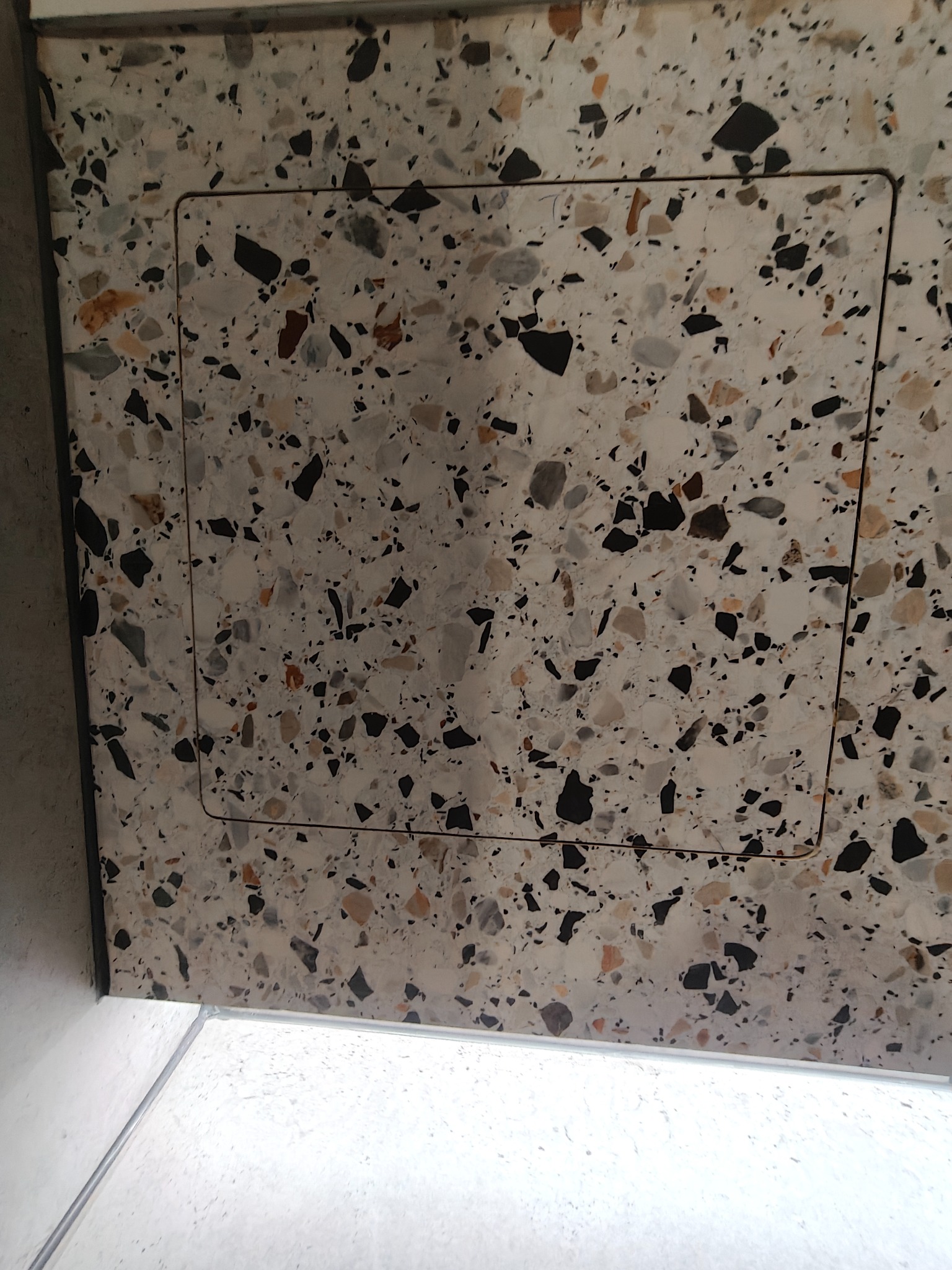 Biała ściana z mozaiką w stylu lastryko, na której widoczny jest ukryty właz rewizyjny z delikatnym obramowaniem. Precyzyjne dopasowanie wzoru.