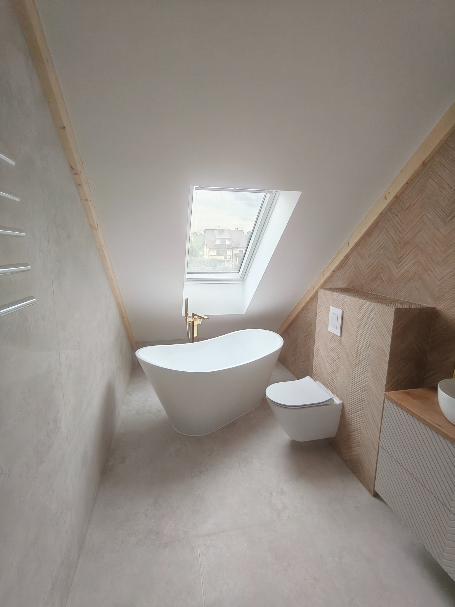 Nowoczesna łazienka na poddaszu z wolnostojącą wanną, wiszącą toaletą i płytkami ułożonymi w jodełkę. Okno dachowe wpuszcza naturalne światło.