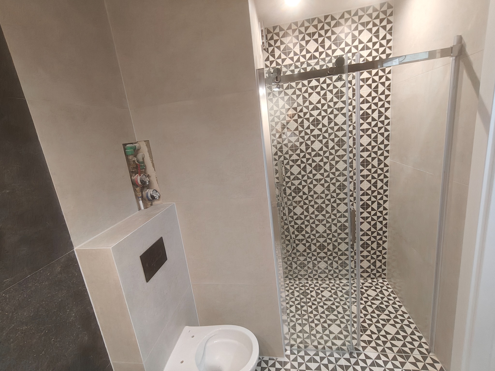Wykończona łazienka z kabiną prysznicową, mozaiką w geometryczne wzory na ścianach i podłodze, oraz podwieszaną toaletą. Widoczne przyłącza wodne.