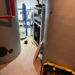 Paweł Hydraulik - Wnętrze kotłowni w trakcie prac: widoczne duże zbiorniki, otwarty piec, narzędzia, instalacja z rur miedzianych i plastikowych oraz ślady wilgoci na podłodze.