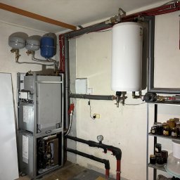 Paweł Hydraulik - Kotłownia z piecem kondensacyjnym, zasobnikiem ciepłej wody, naczyniami przeponowymi oraz instalacją grzewczą i hydrauliczną, widoczne rury w izolacji, manometr oraz regał z przetworami.