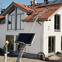 Paweł Hydraulik - Instalacja paneli słonecznych na dachu domu jednorodzinnego z użyciem specjalistycznej windy dekarskiej; pracownicy w kaskach na dachu, operator windy w żółtym kasku na ziemi.