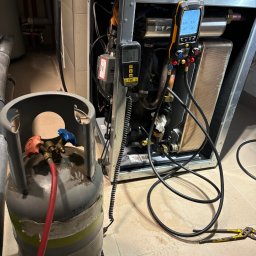 Paweł Hydraulik - Podłączony cylinder z czynnikiem chłodniczym do urządzenia chłodniczego z widocznymi manometrami cyfrowymi wskazującymi ciśnienie, w tle rury instalacji i płytki podłogowe.