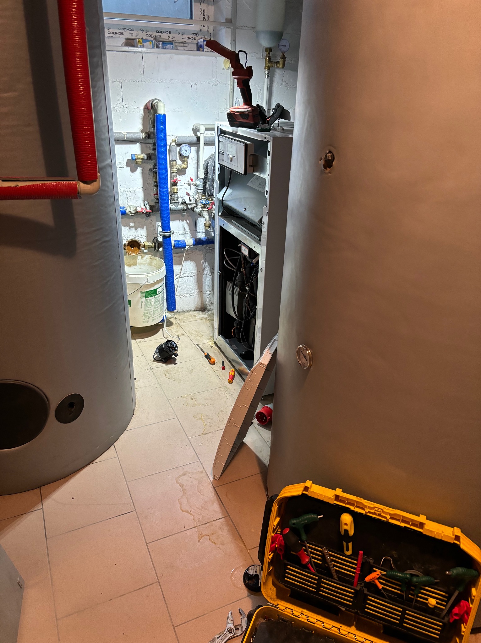 Wnętrze kotłowni w trakcie prac: widoczne duże zbiorniki, otwarty piec, narzędzia, instalacja z rur miedzianych i plastikowych oraz ślady wilgoci na podłodze.