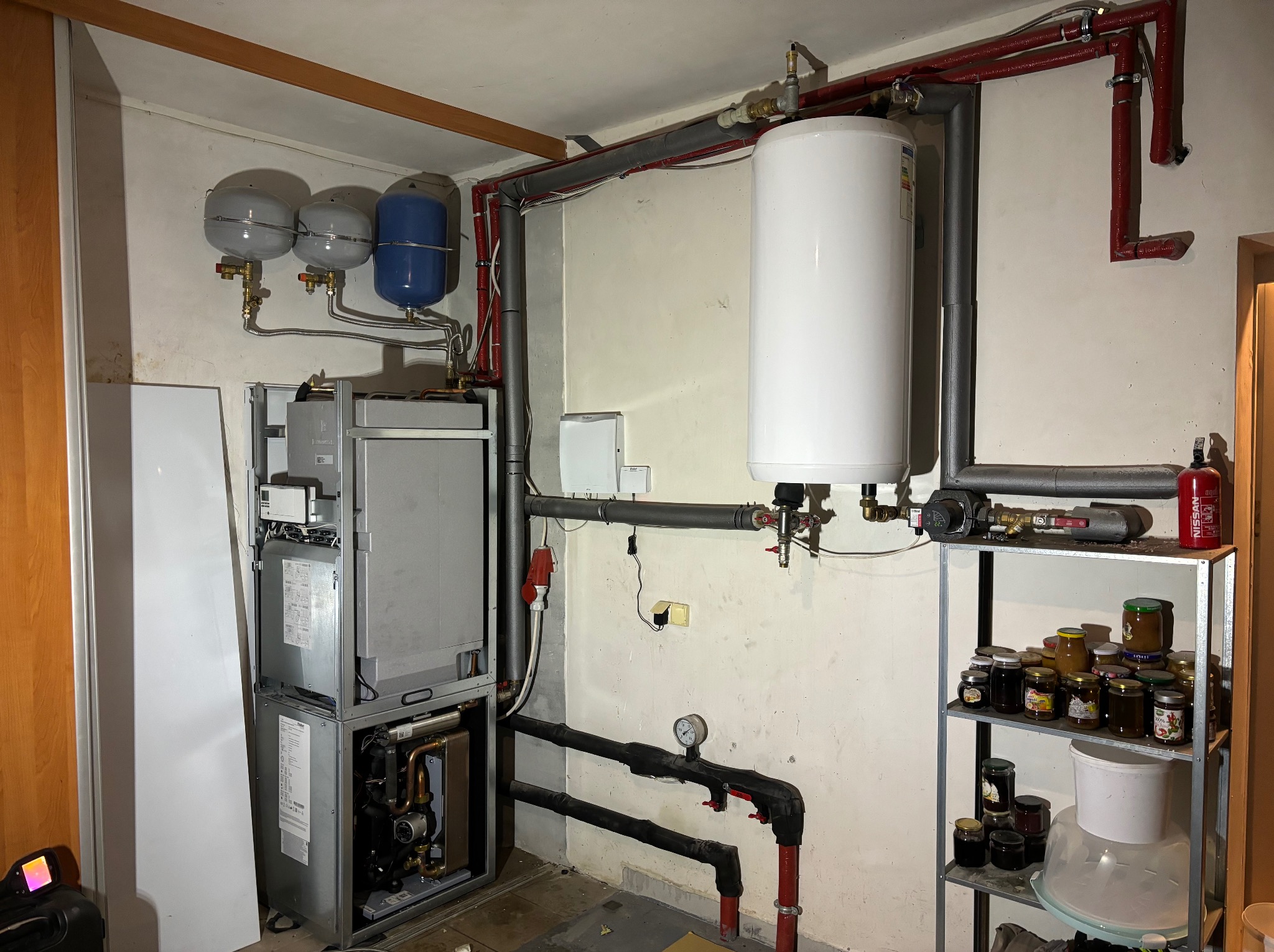 Kotłownia z piecem kondensacyjnym, zasobnikiem ciepłej wody, naczyniami przeponowymi oraz instalacją grzewczą i hydrauliczną, widoczne rury w izolacji, manometr oraz regał z przetworami.